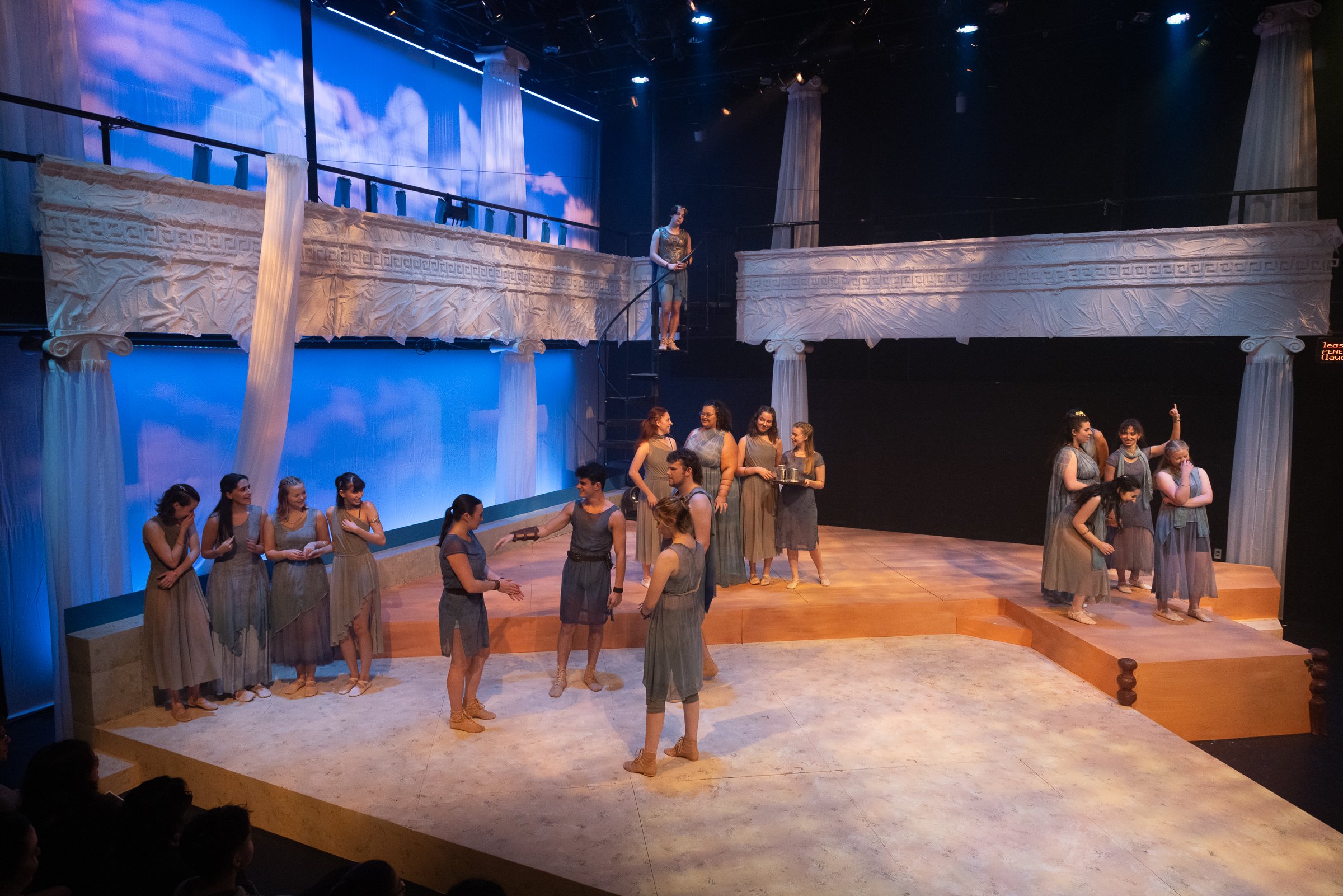The stage set resembles ancient Greek architecture with white columns and drapes, and a backdrop of a blue sky with clouds. Several actors are on stage, some engaging in conversation and others standing in groups.