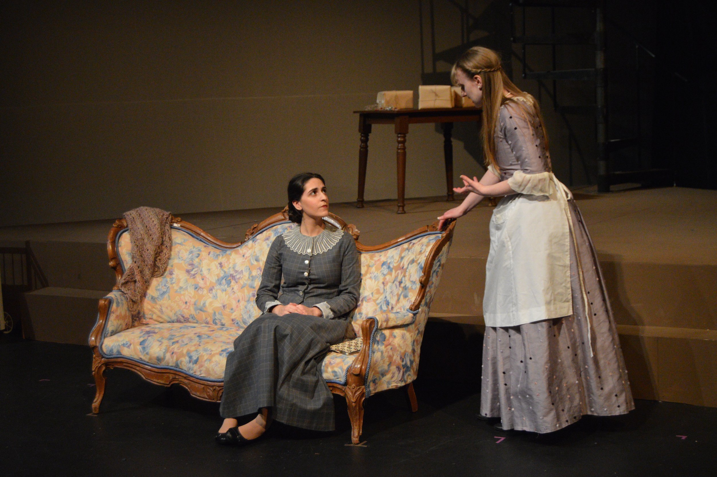 Two women performing in a theater; one seated on a vintage floral sofa and the other standing and engaging her in conversation, dressed in historical costumes.