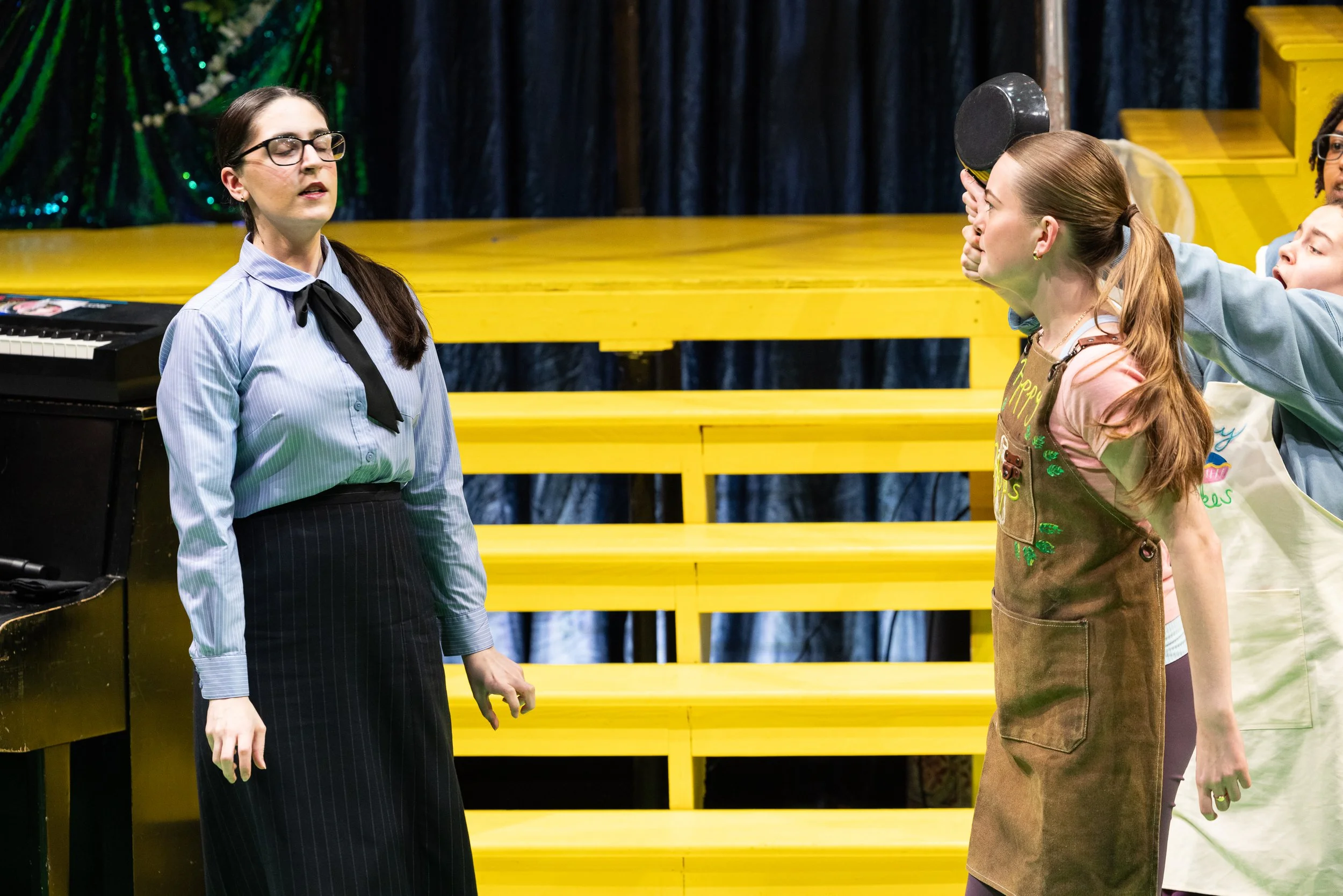 Three young women on a stage. One with glasses and a blue shirt stands with her eyes closed. The others, with reddish hair, hold objects to their eyes as if using mirrors. The background includes a yellow bench and dark curtains.