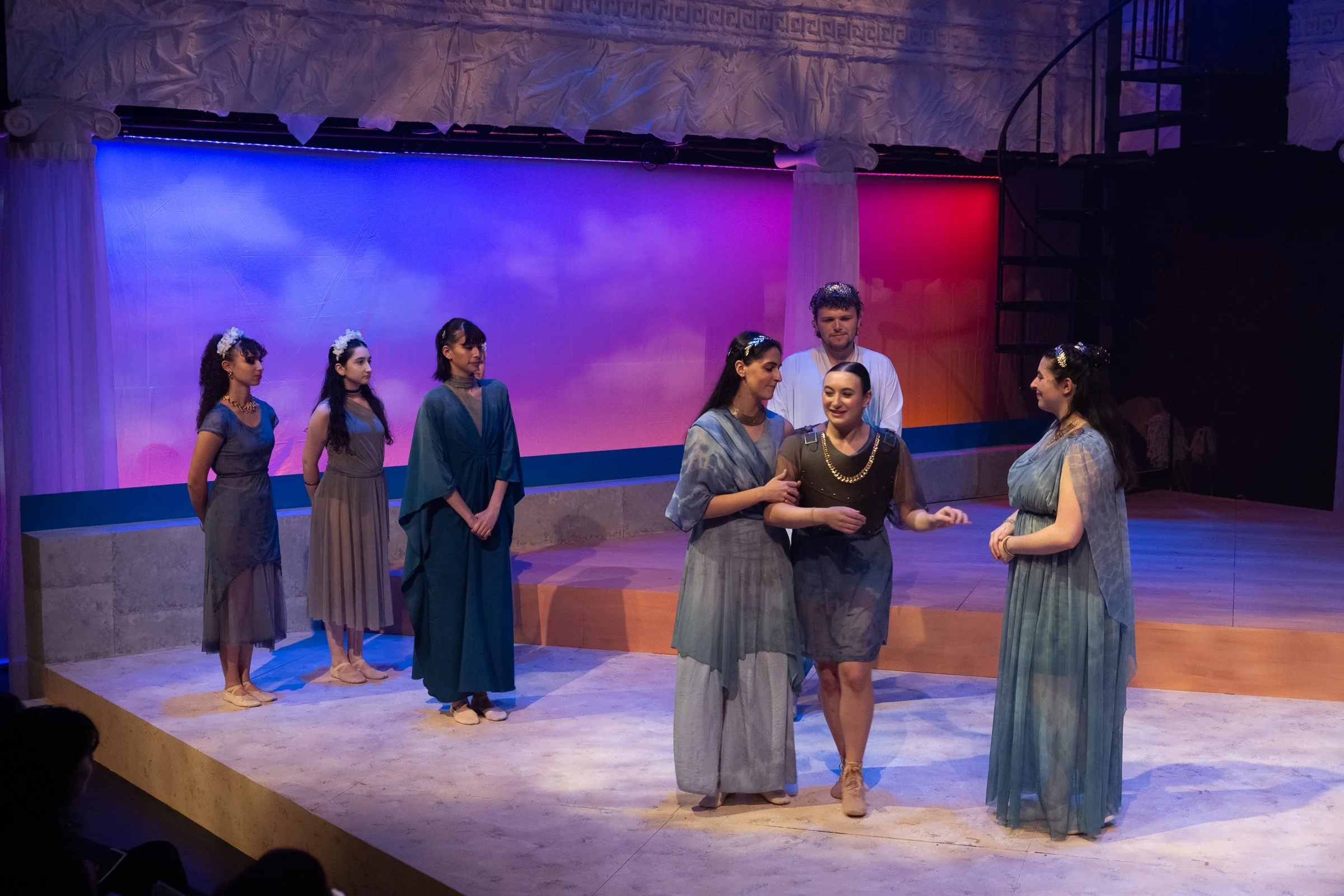 A group of eight actors performing on stage with a colorful backdrop. The actors are dressed in costumes resembling ancient Greek attire, and they are engaged in a scene, with some talking and others listening.