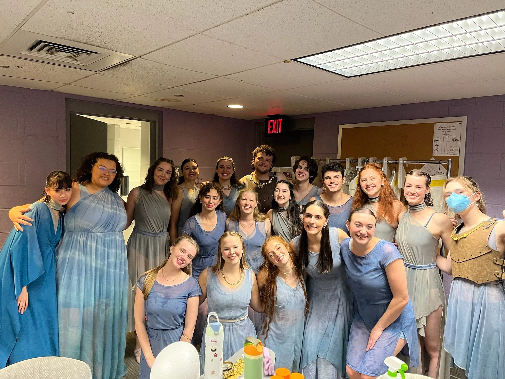 A group of young people dressed in various shades of blue and gray, smiling for a group photo in an indoor setting with purple walls and a ceiling with fluorescent lights. Some are holding bottles and water bottles are on the table in front of them.