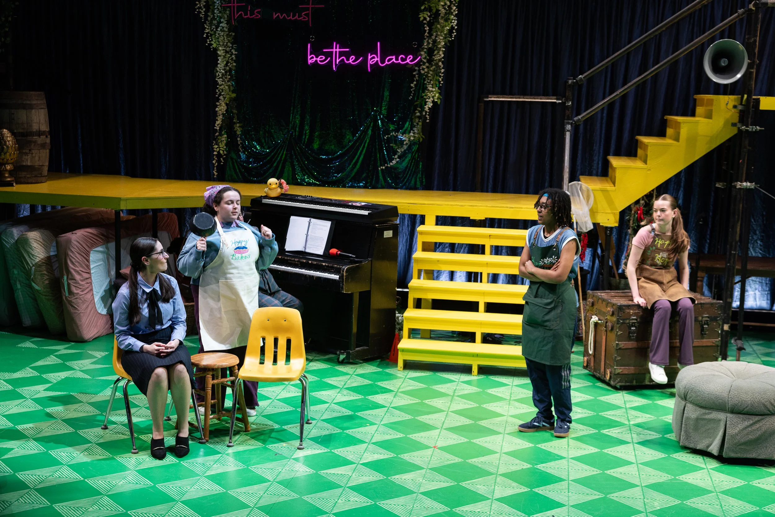 The image shows a stage with a neon sign that says 'this must be the place,' decorated with greenery and white flowers. There are four people in front of the stage, one sitting while three stand, and a piano with a rubber duck on it. The setting appe