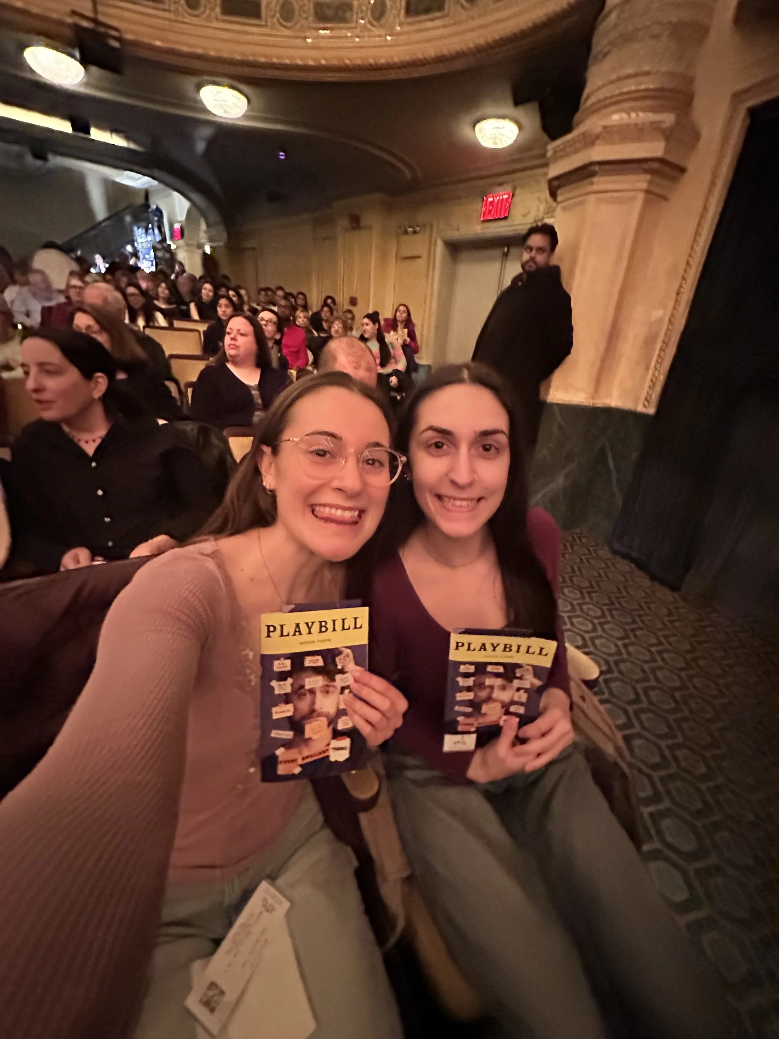 Two young women sitting in a theater, smiling and holding Playbill programs, with a large audience and ornate theater interior in the background.