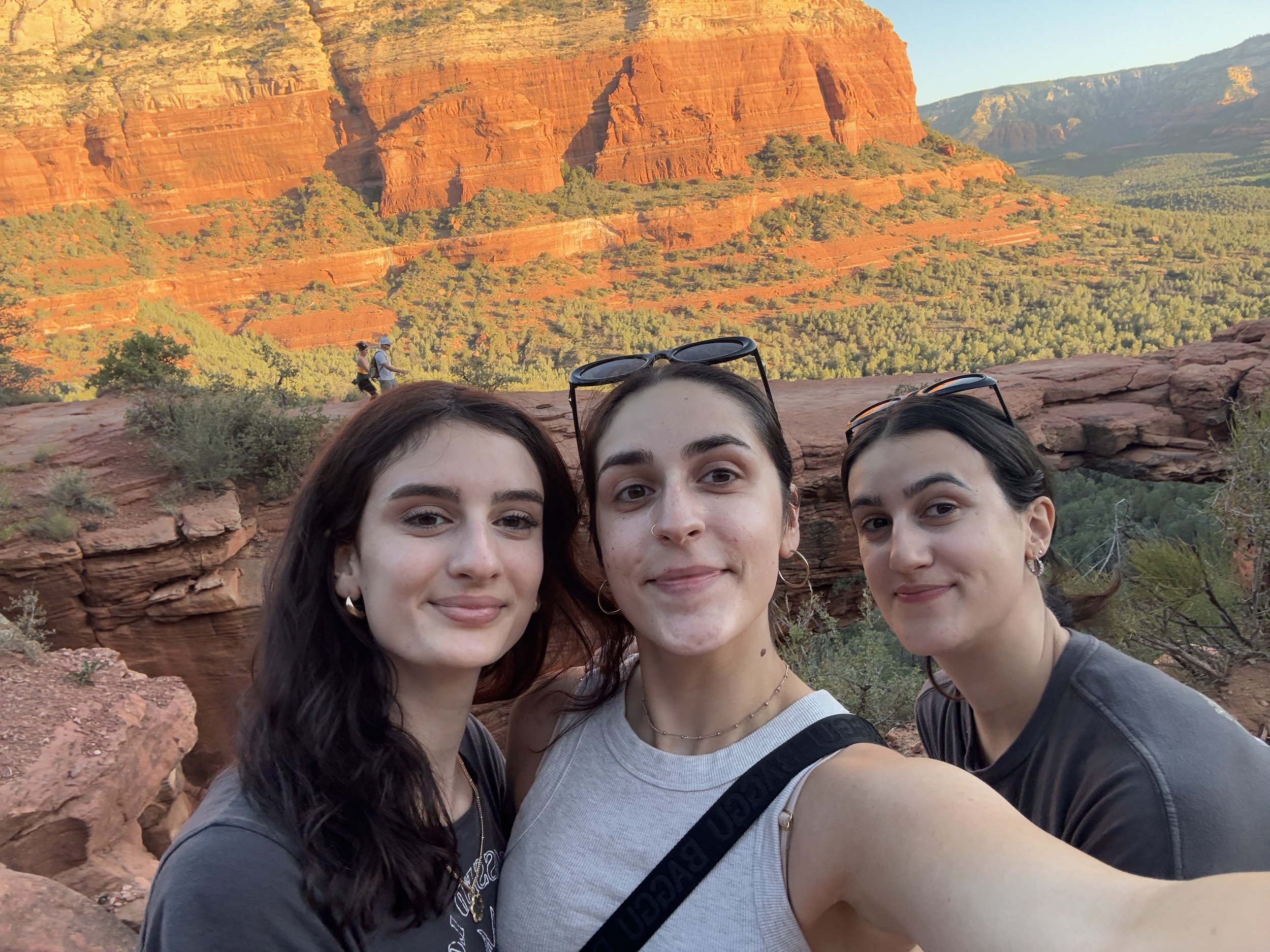 A group of three young women taking a selfie with a red rock canyon landscape in the background, featuring towering red cliffs and green vegetation.