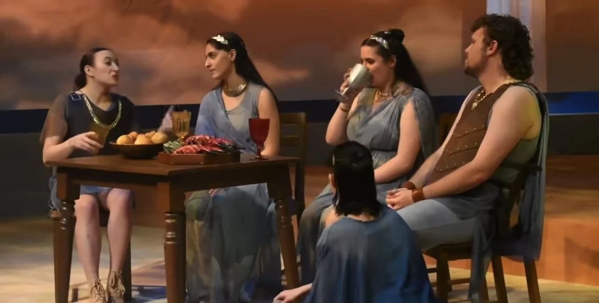 Cast of five people in ancient Greek costumes seated around a table with food and drinks, in a theatrical setting.