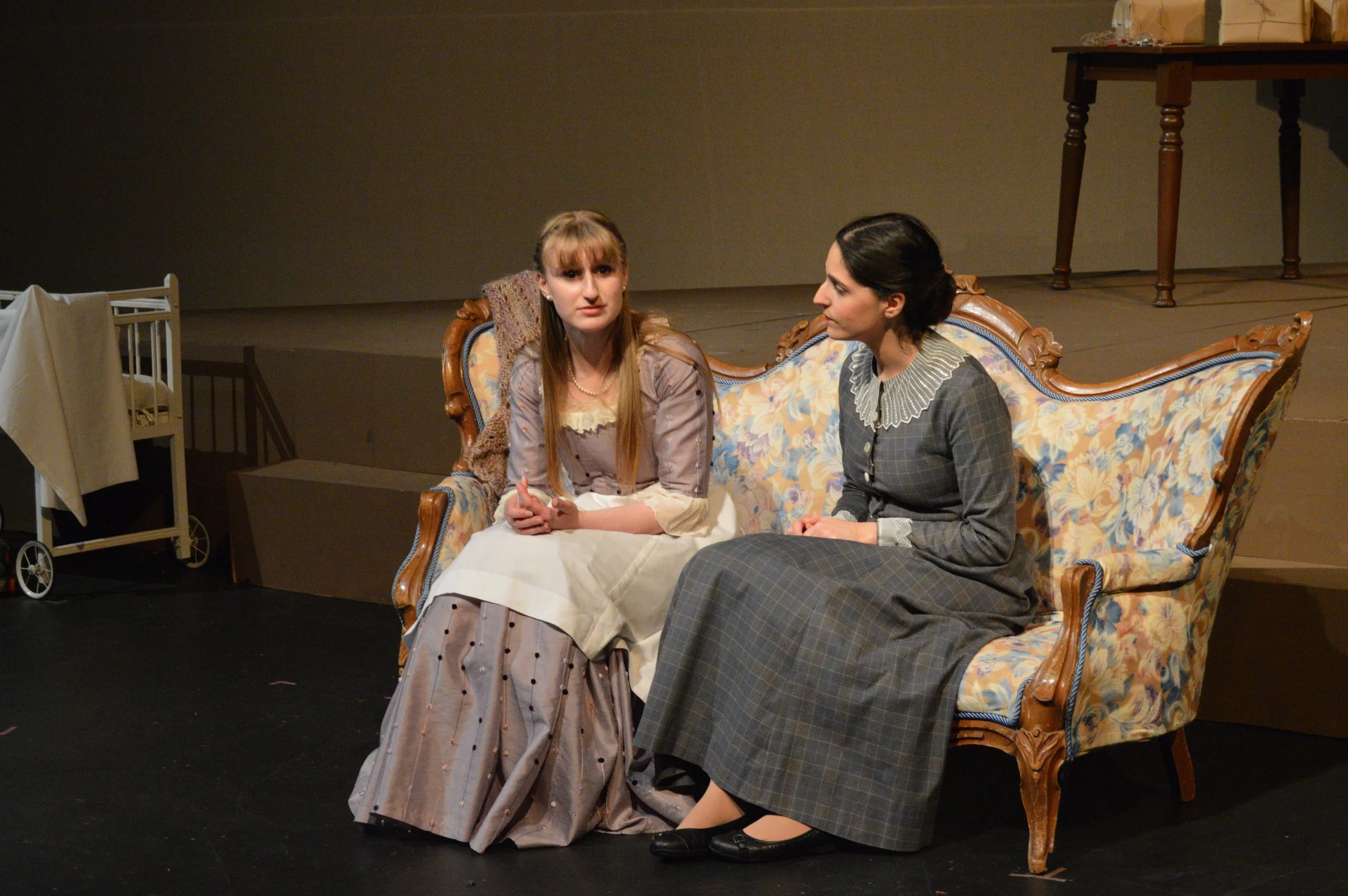 Two women sitting on a vintage floral sofa, engaged in a conversation on a stage with minimal background and props.