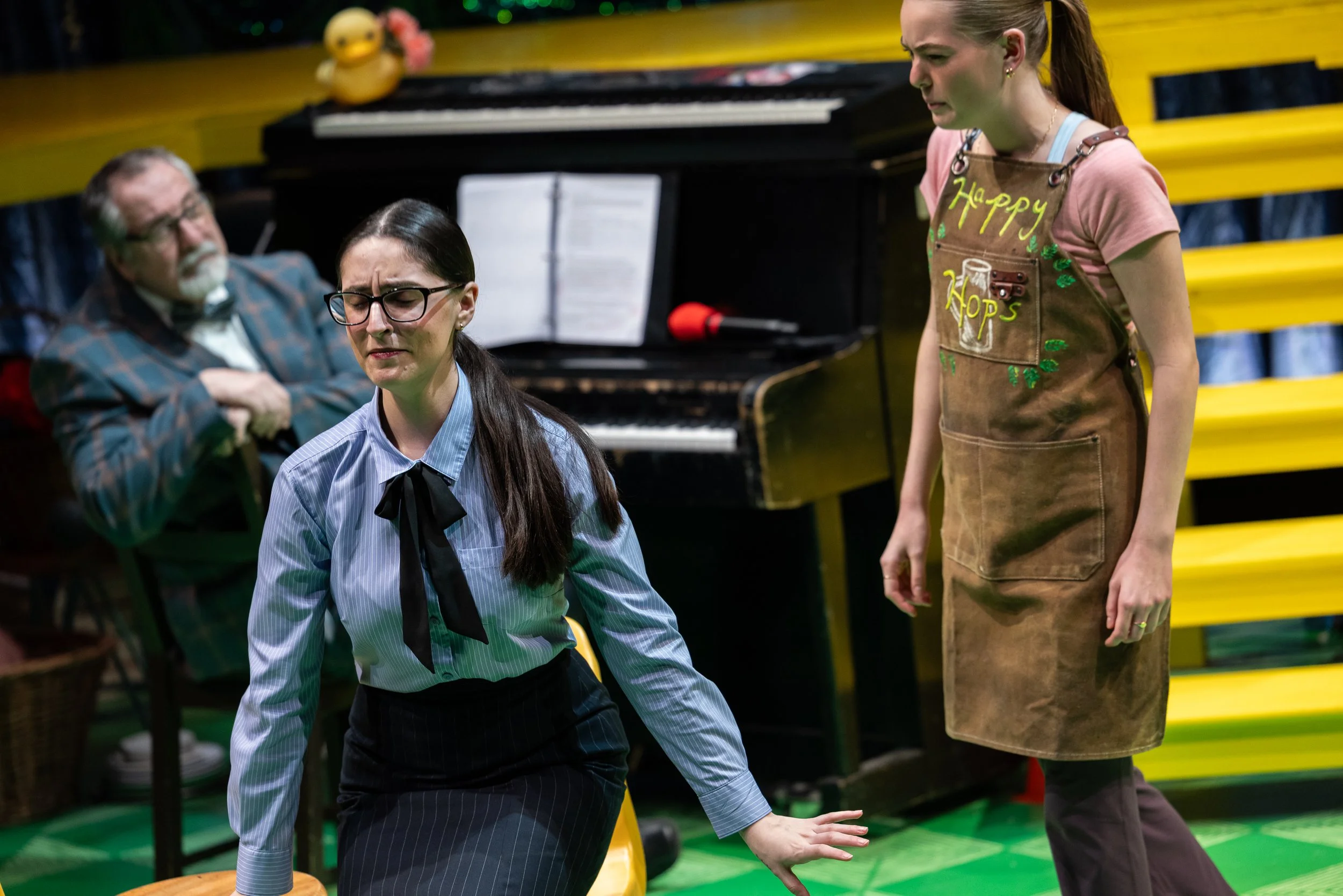 A woman with glasses and a striped shirt moves on her hands and knees, while another woman standing nearby appears to be talking or instructing. In the background, an older man with glasses and a beard sits with his arms crossed. The scene takes plac
