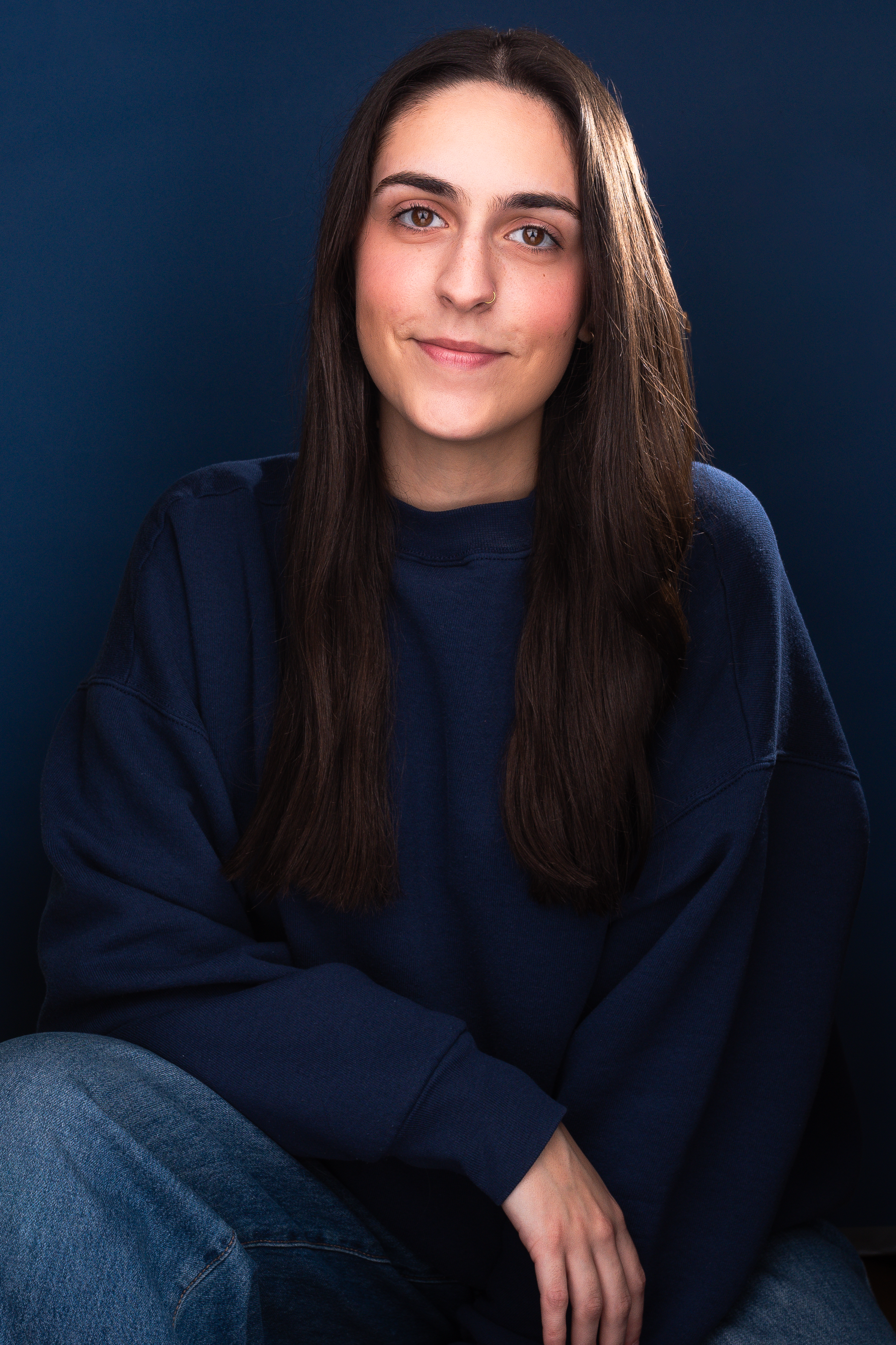 A young woman with long dark hair, light skin, and brown eyes, wearing a navy blue sweatshirt, sitting against a dark blue background.