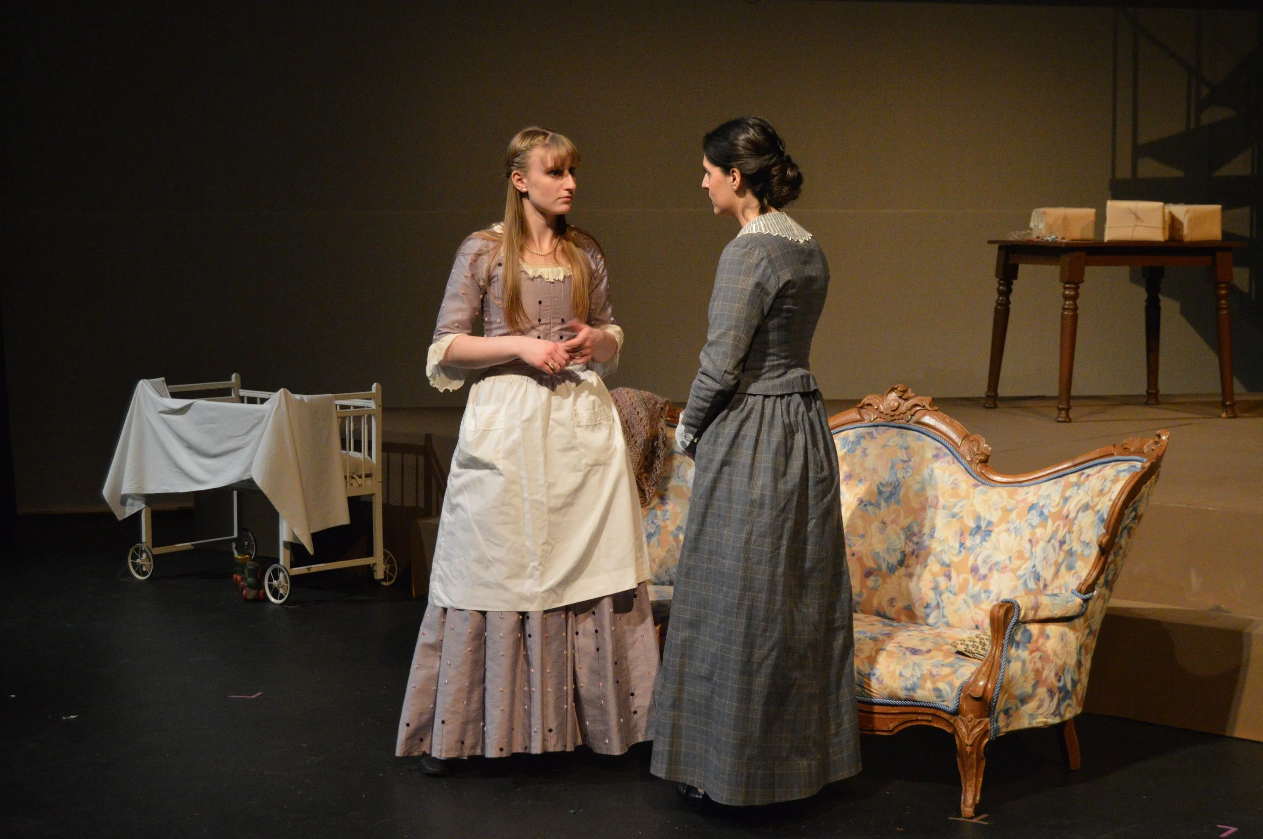 Two women in period costumes are standing face to face on a stage with a set that includes a crib, a sofa, and a table with wrapped packages.