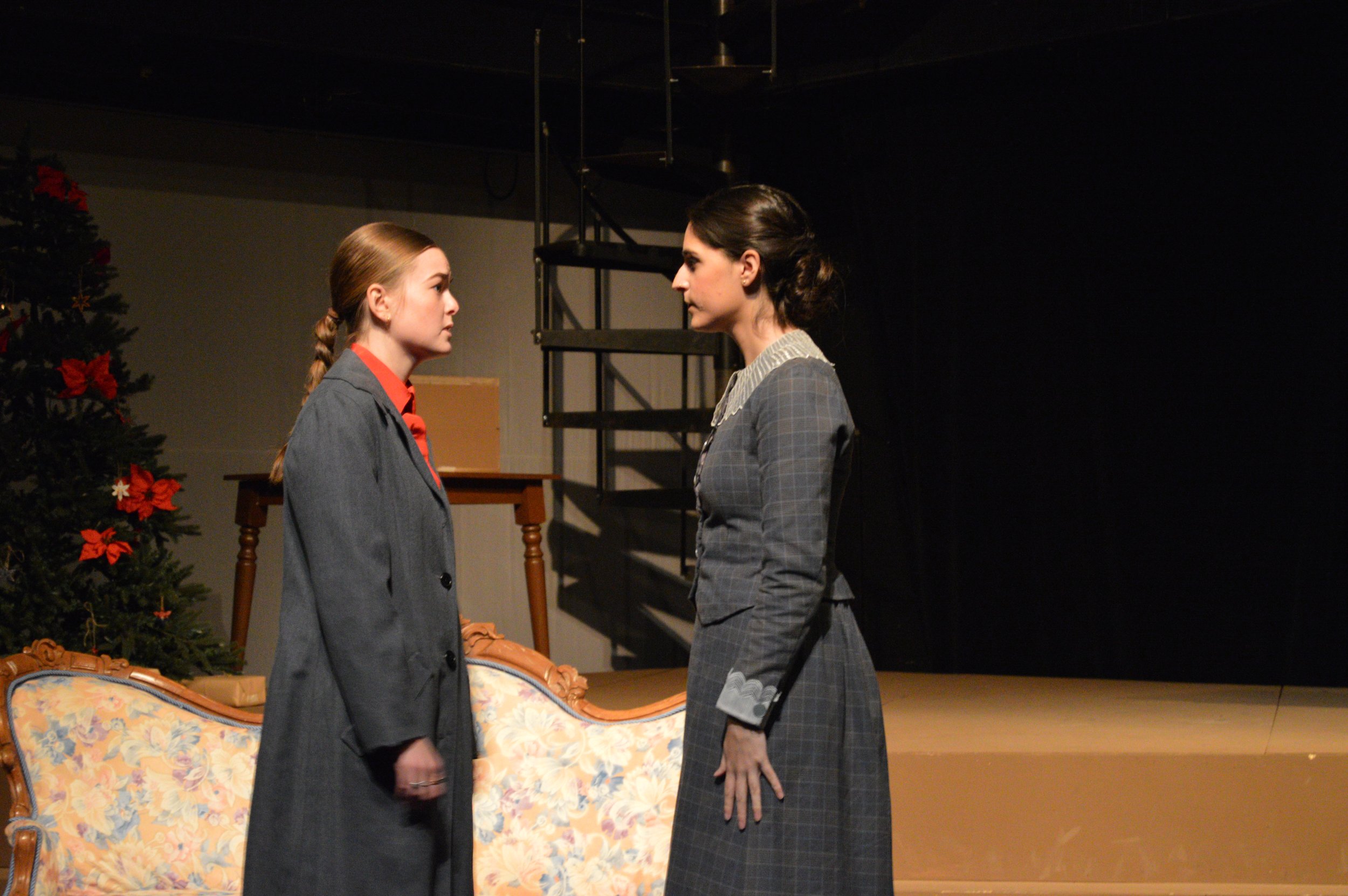 Two women standing face to face on stage in a theater, with a decorated Christmas tree visible on the left side, a sofa in the foreground, and a black background. They are dressed in vintage-style gray suits and appear to be engaged in a serious conv