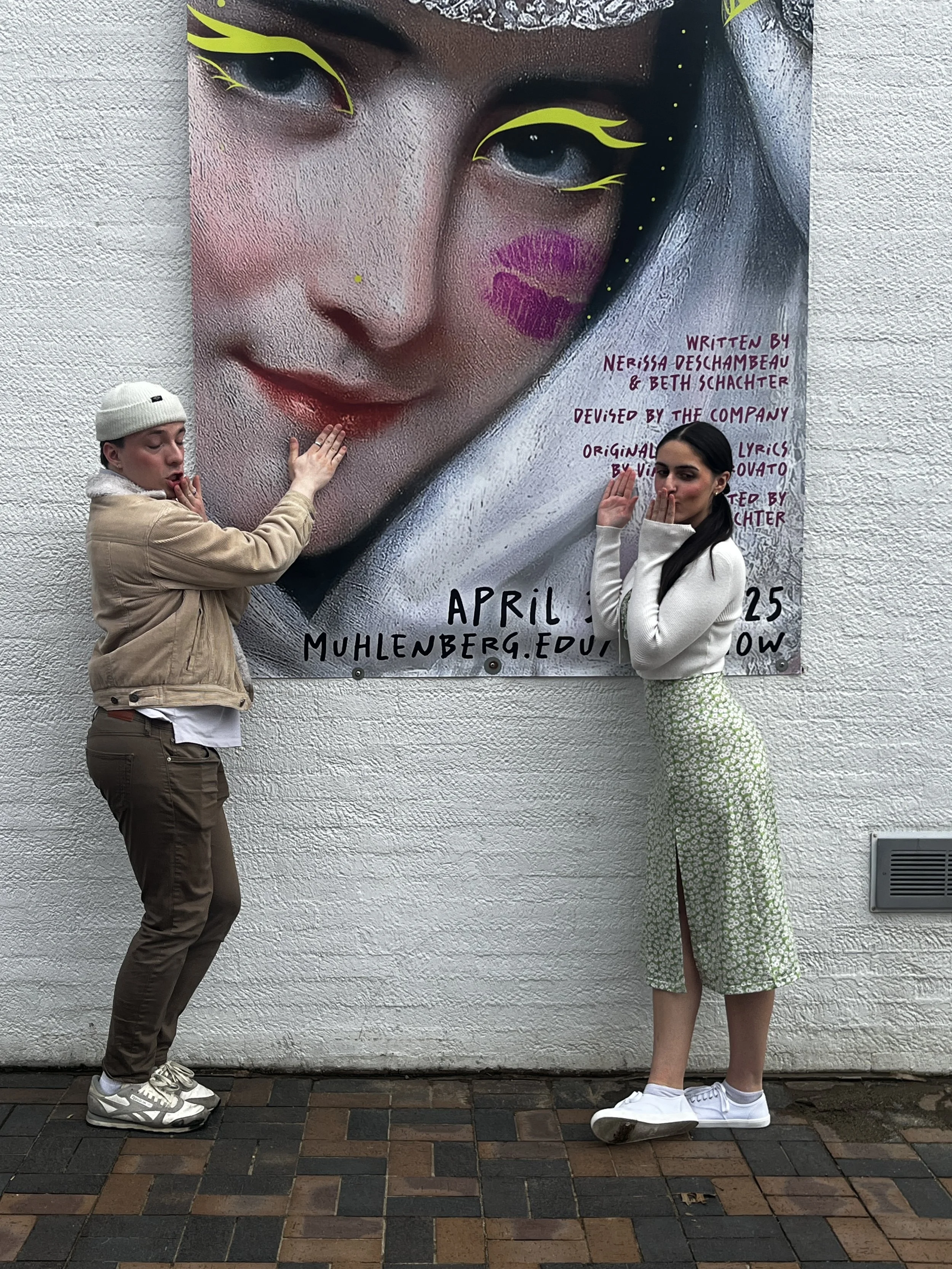 Two people pose in front of a large poster of a woman's face, with one person on each side. The poster features a woman with makeup, lipstick, and an artistic design around her eyes. The person on the left is wearing a beige jacket, brown pants, gray