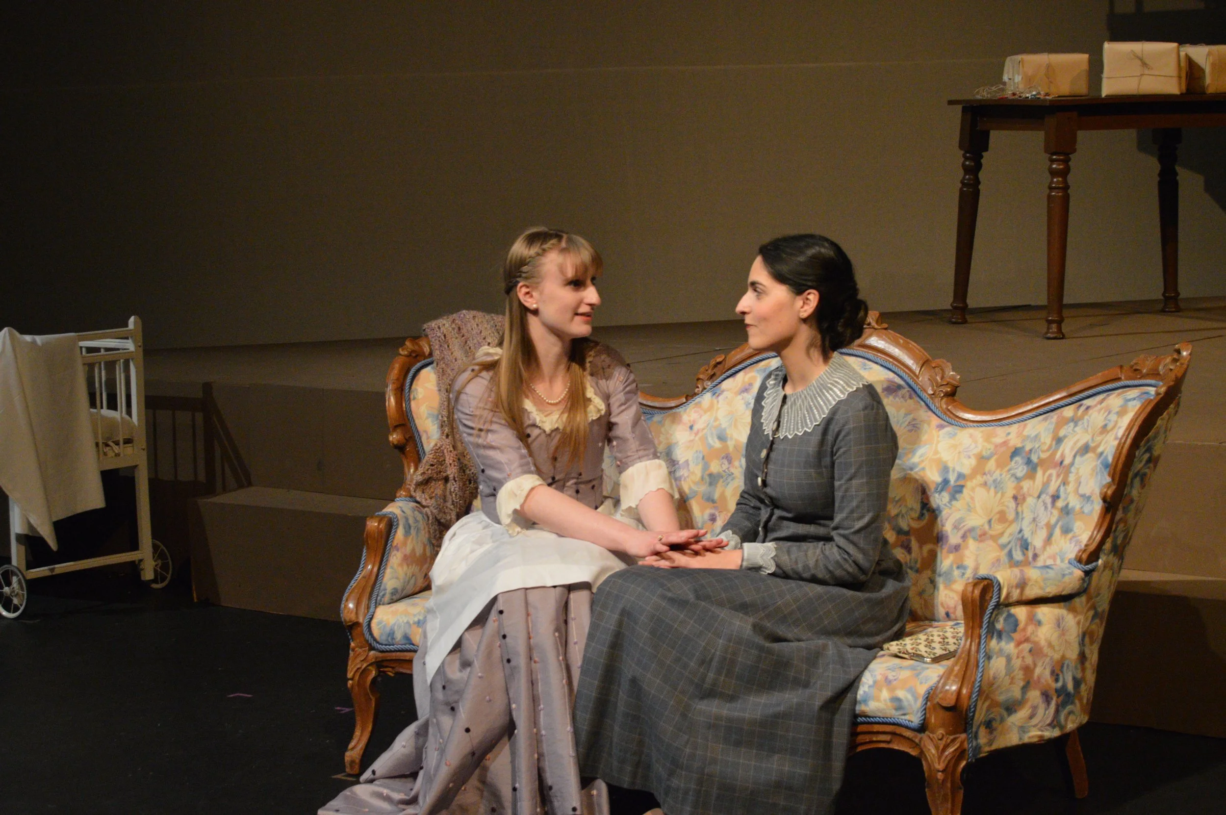 Two women dressed in vintage clothing sitting on a floral-patterned sofa and holding hands, engaged in conversation. The setting appears to be a theatrical stage with a plain backdrop and minimal furniture, including a small side table and a crib.
