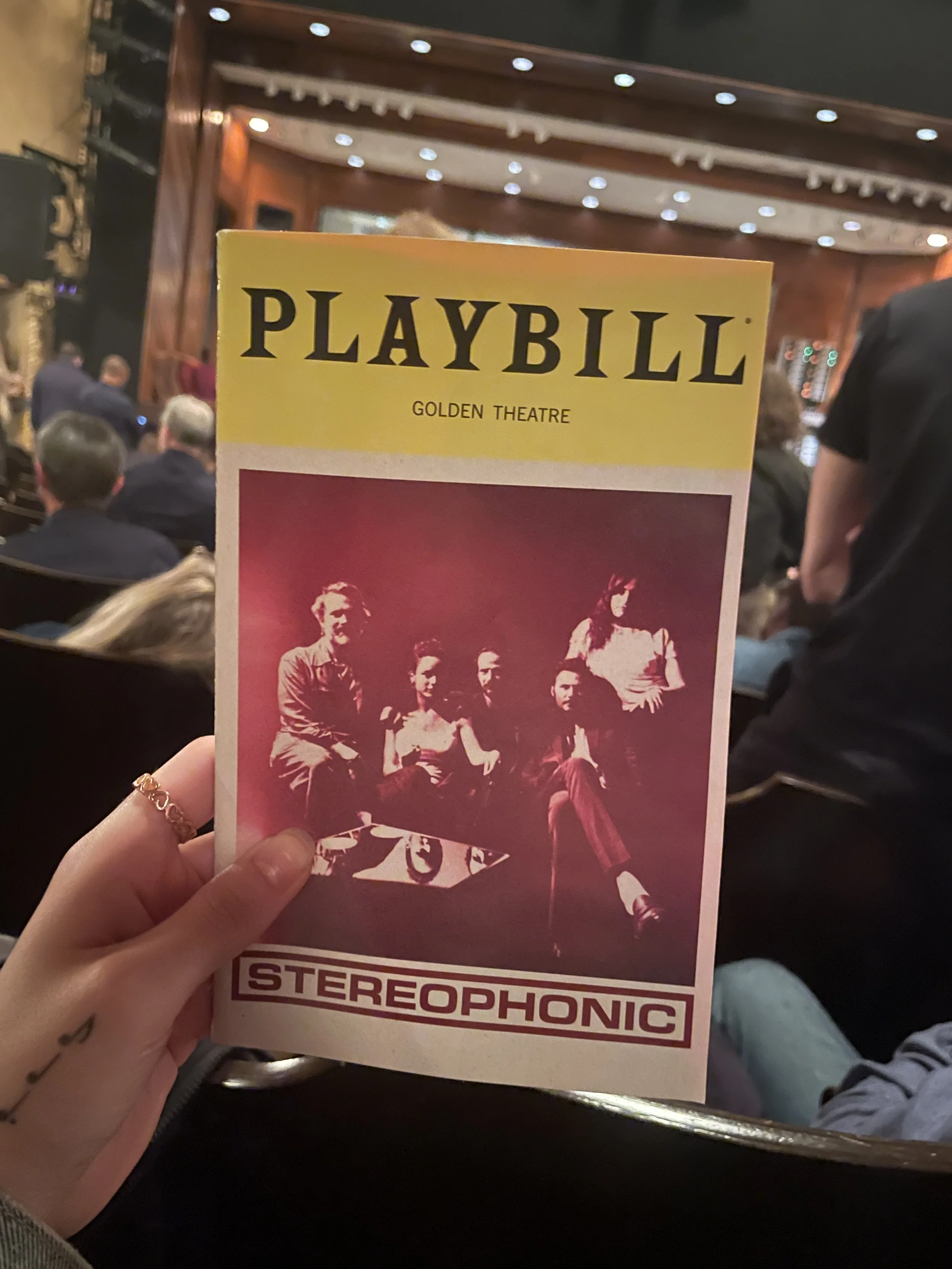 A person holding a Playbill theater program for the musical 'STEREOPHONIC' at the Golden Theatre, with the theater's interior and audience in the background.