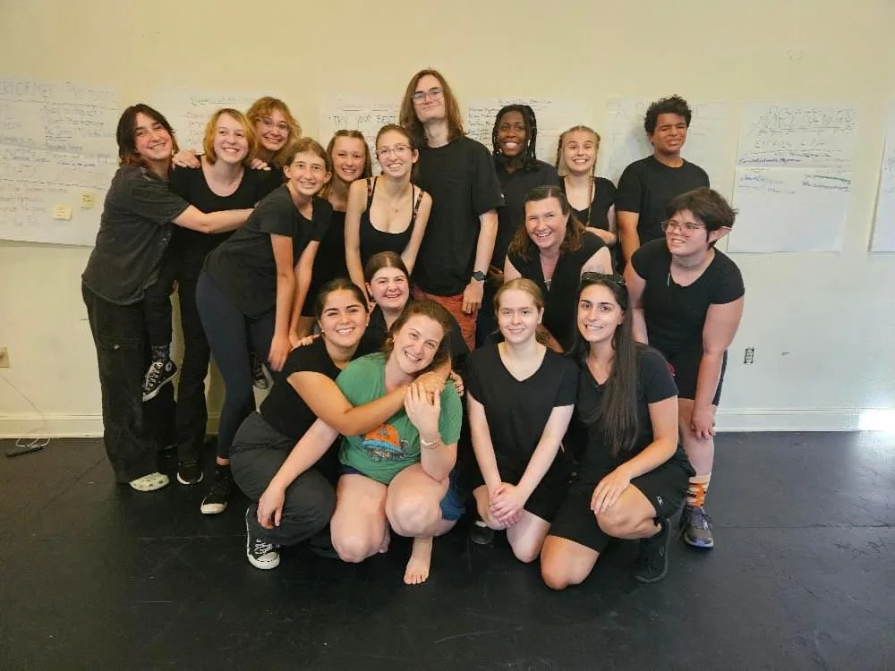 Group of 14 young people, mostly women, posing together indoors against a white wall with handwritten posters, all wearing black shirts, smiling and showing camaraderie.