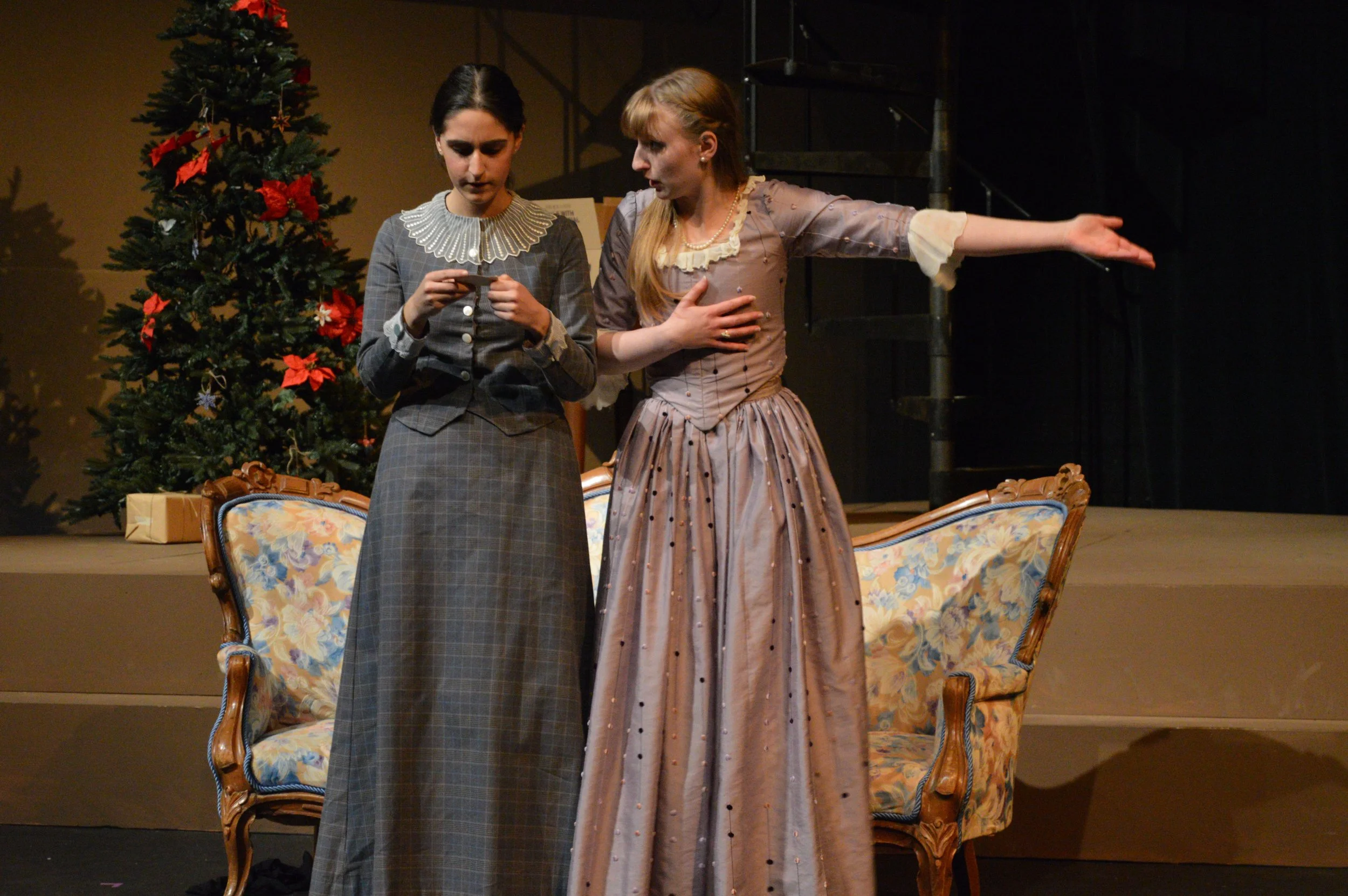 Two women dressed in vintage-style clothing appear to be acting or performing on stage. One is looking at a small piece of paper, and the other is speaking or singing, gesturing with her arm extended. Behind them, there is a decorated Christmas tree 