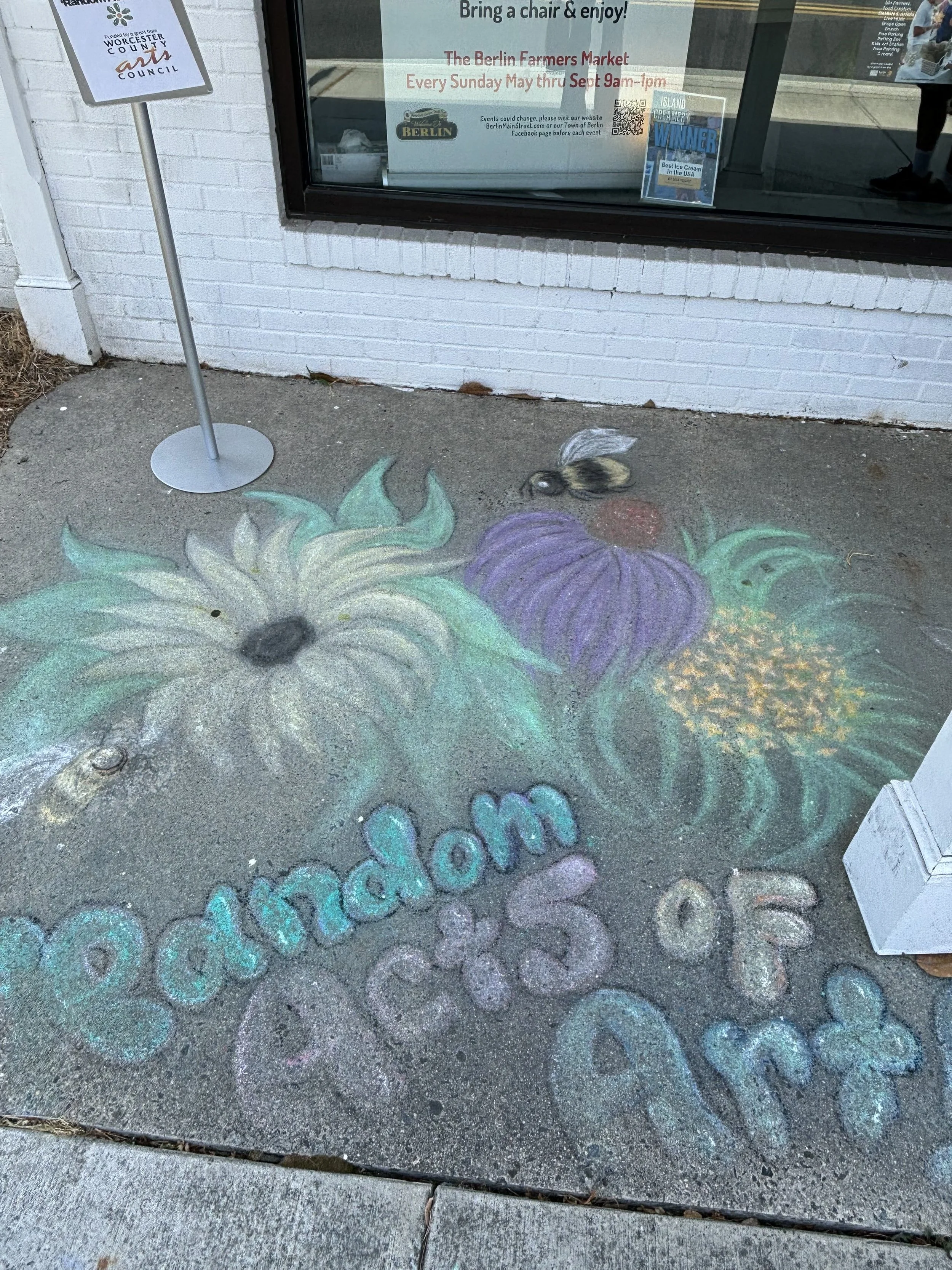 Colorful chalk art on a concrete sidewalk depicting sunflowers, a bee, purple and green plants, and the word 'welcome' in multicolored letters.