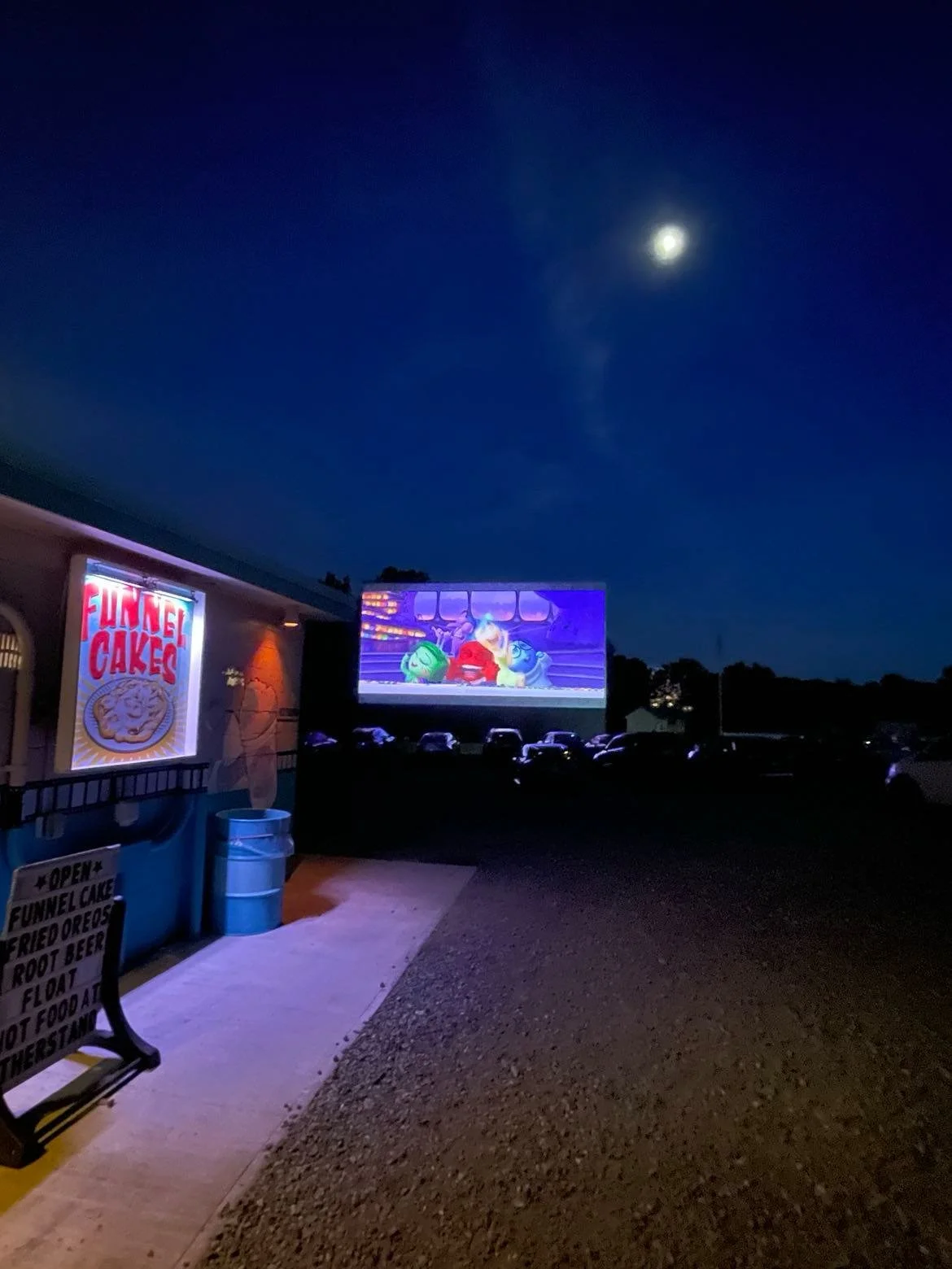 Outdoor drive-in theater showing animated movie at night, with illuminated sign for funnel cakes and a partly cloudy moon in the sky