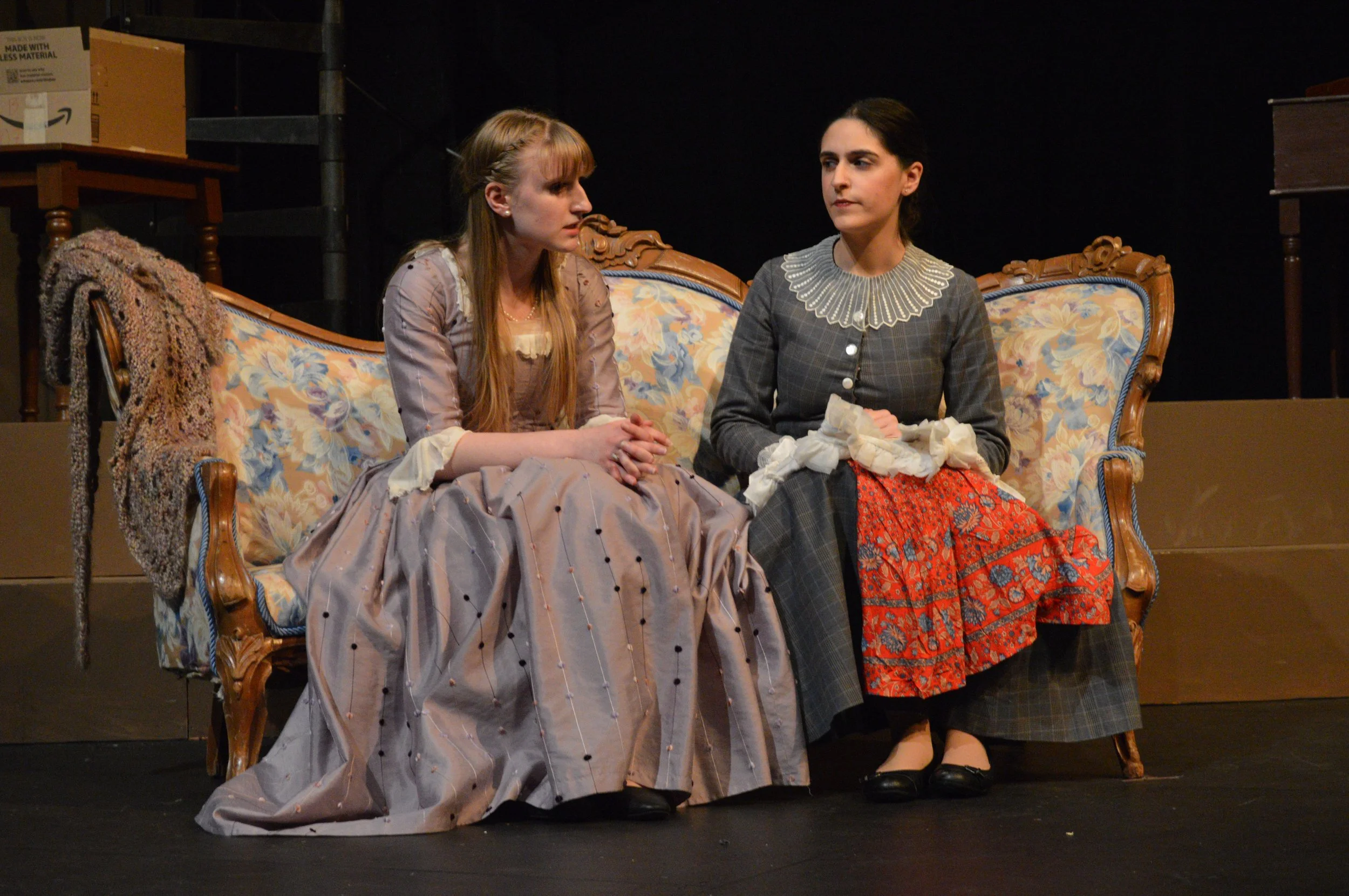Two women in period costumes sit on a floral upholstered settee on stage during a theatrical performance. One woman is speaking while the other appears to listen. The set includes a blanket draped over the arm of the settee and cardboard boxes in the