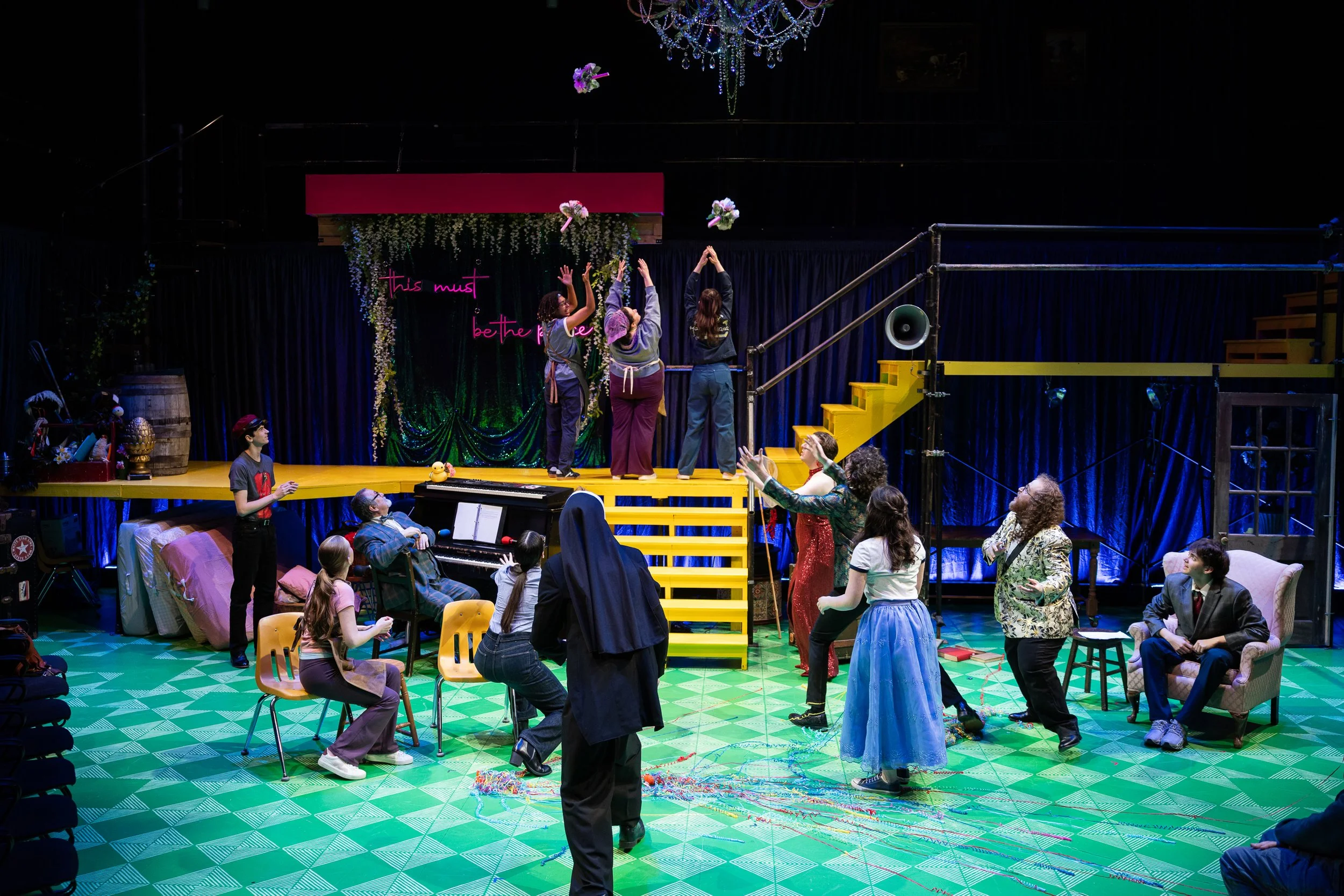 Theater stage with performers and audience, decorated with flowers, colorful lights, and a neon sign reading 'this must be the place.' Audience members are seated and engaging with the performance while some actors interact on stage.