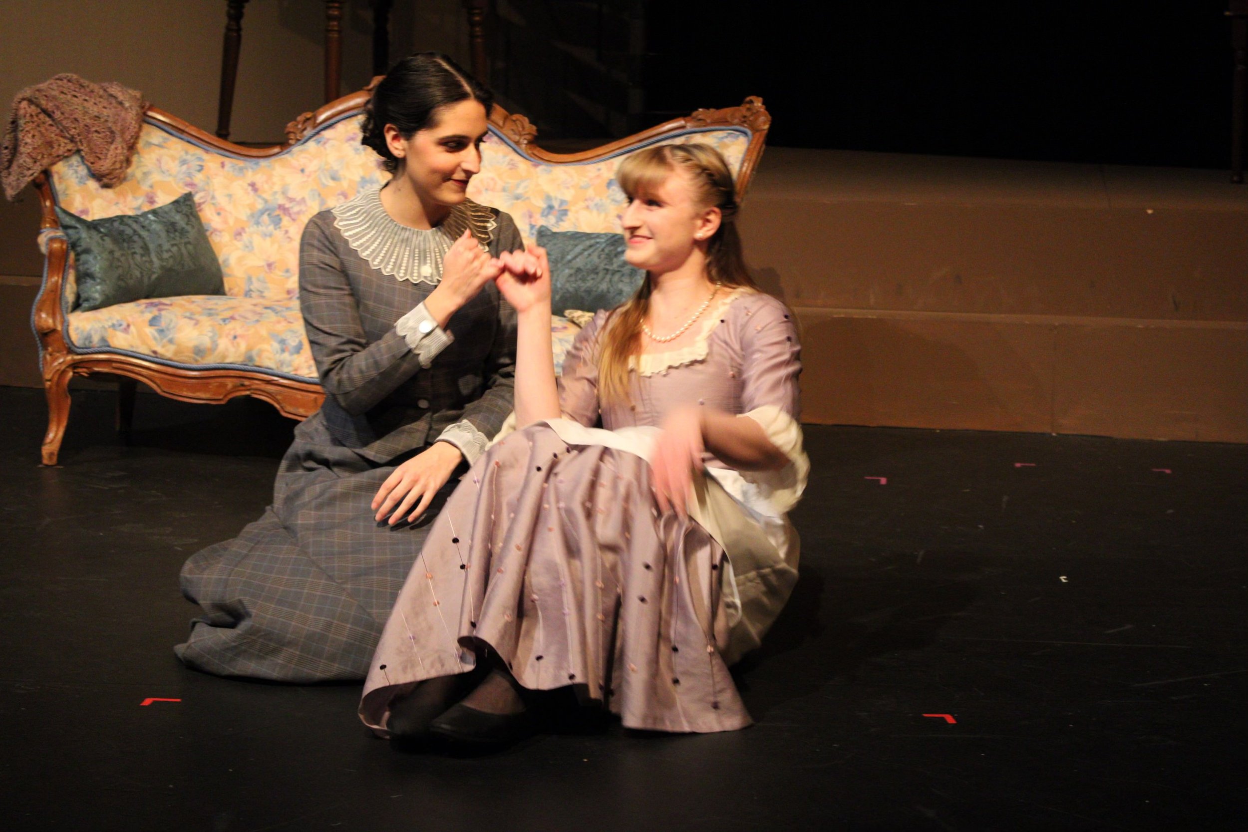 Two women are sitting on the stage floor, holding hands with fingers intertwined, looking at each other and smiling. The woman on the left has dark hair and is wearing a checked dress with a detailed collar. The woman on the right has blonde hair and
