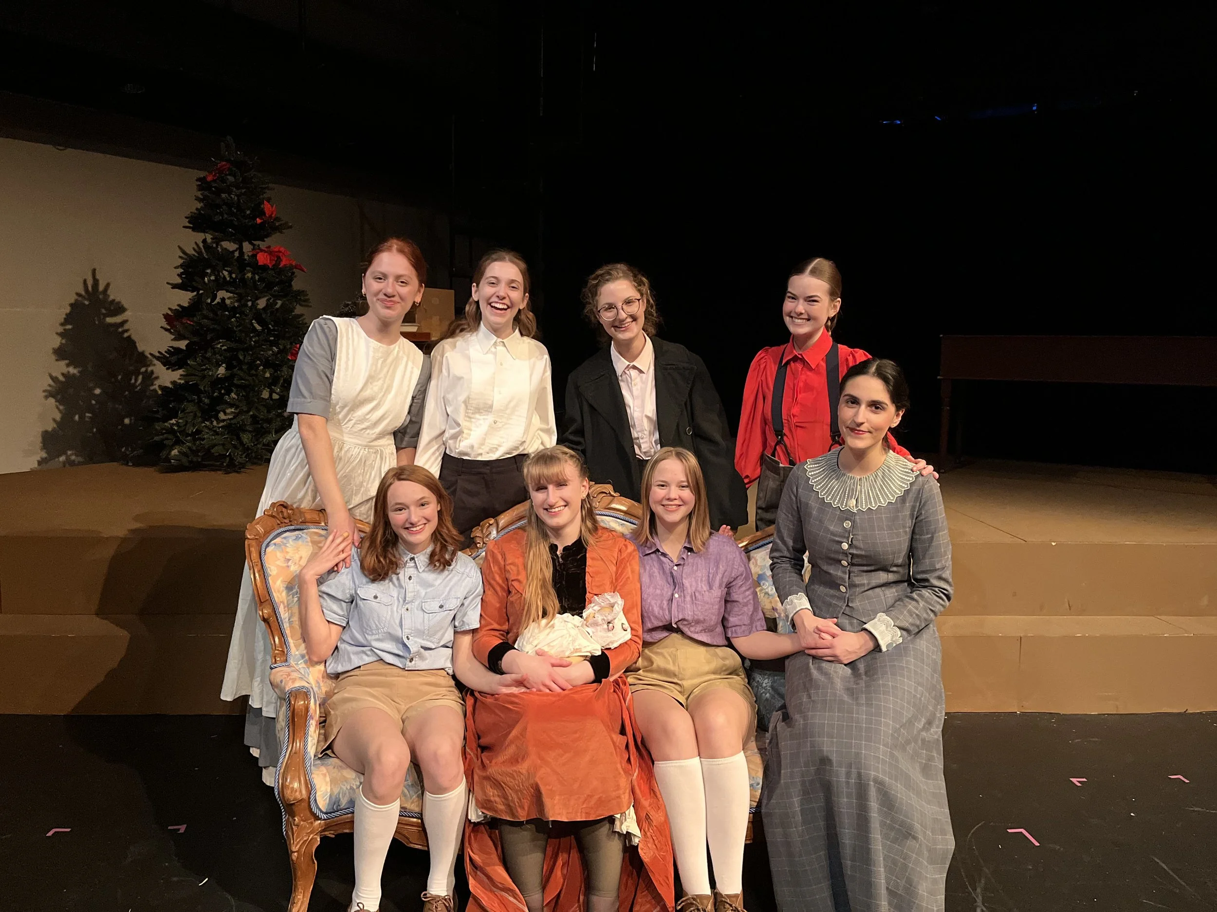 Group of young women in vintage and period costumes posing on stage with a Christmas tree in the background.