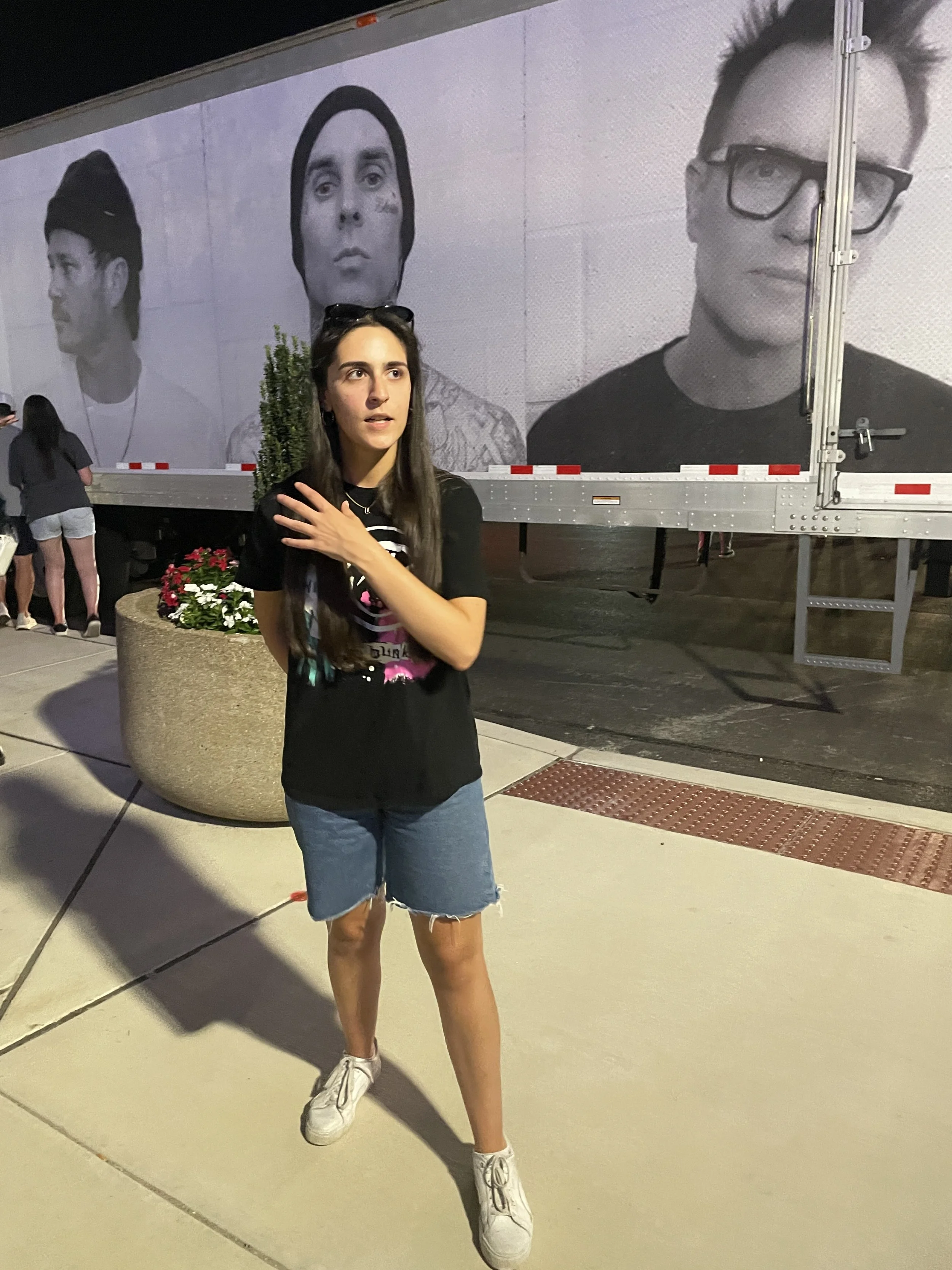 A woman with long dark hair, wearing a black t-shirt, denim shorts, and white sneakers, standing outside at night. She has sunglasses resting on her head and appears to be talking or reacting to something. Behind her is a large trailer with black and