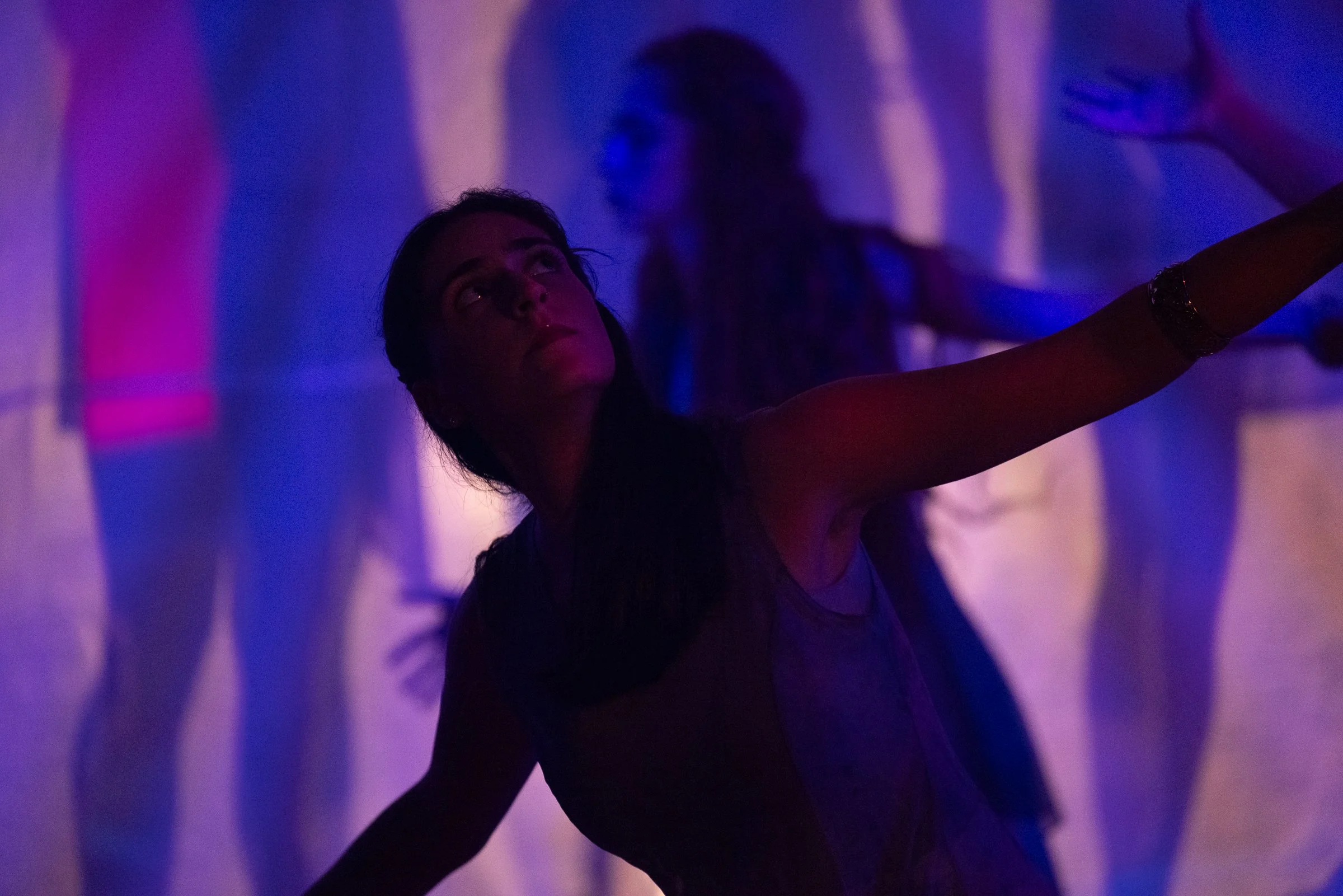 A woman with dark hair in a dimly lit room with purple and blue lighting, taking a selfie with her arm extended.