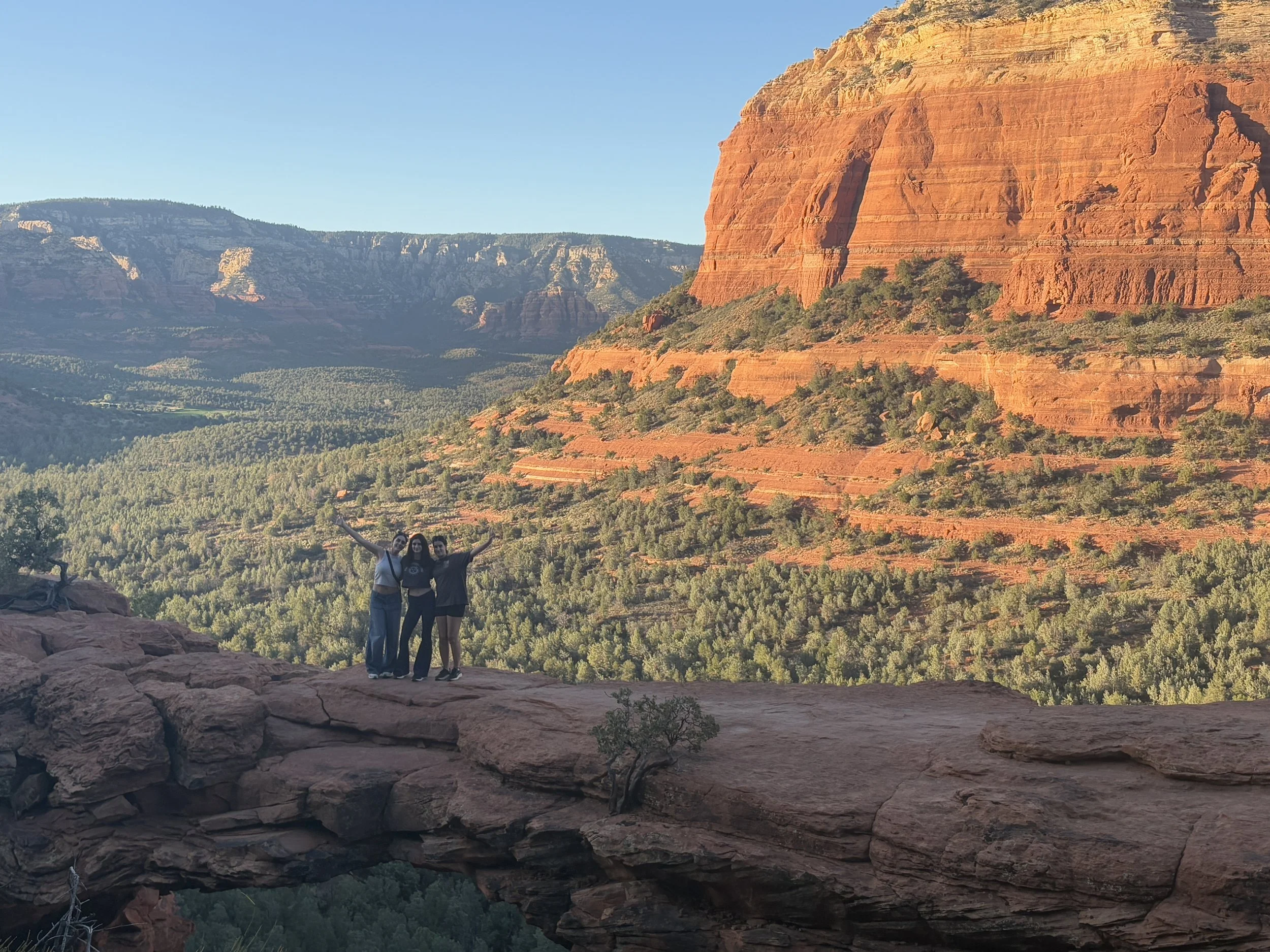 Three people standing on a rock ledge in front of red rock formations and a green valley with trees, under a clear blue sky.