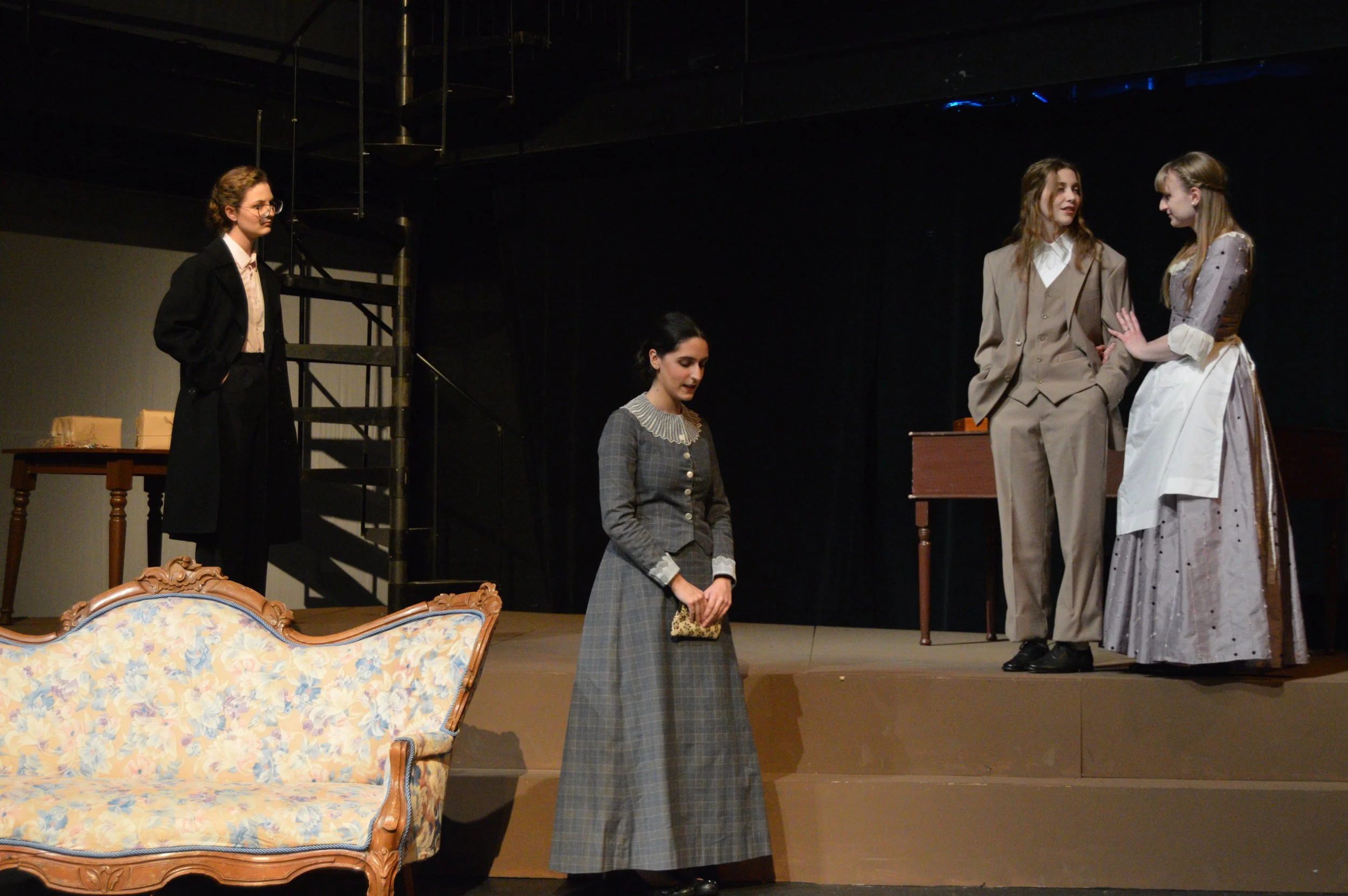 Scene from a theatrical play with four women on stage dressed in vintage clothing, with furniture and a black backdrop.