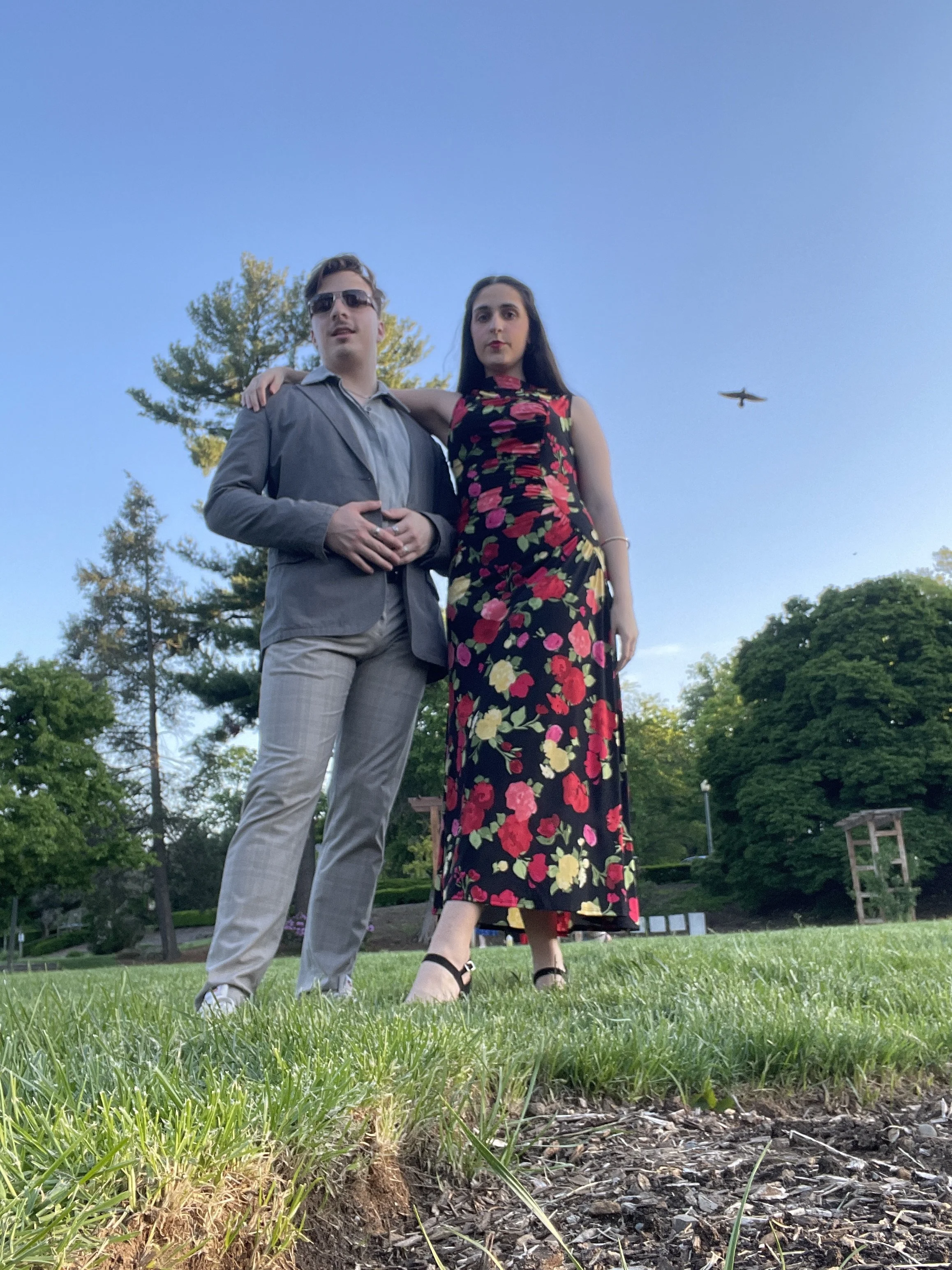 A young man wearing sunglasses and a young woman in a black floral dress stand closely together outdoors on a grassy area, with trees and a blue sky in the background.