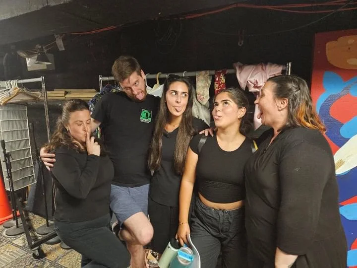 Group of five young women and one young man posing together indoors, some making playful gestures, with a backdrop of colorful abstract wall art and clothing racks.