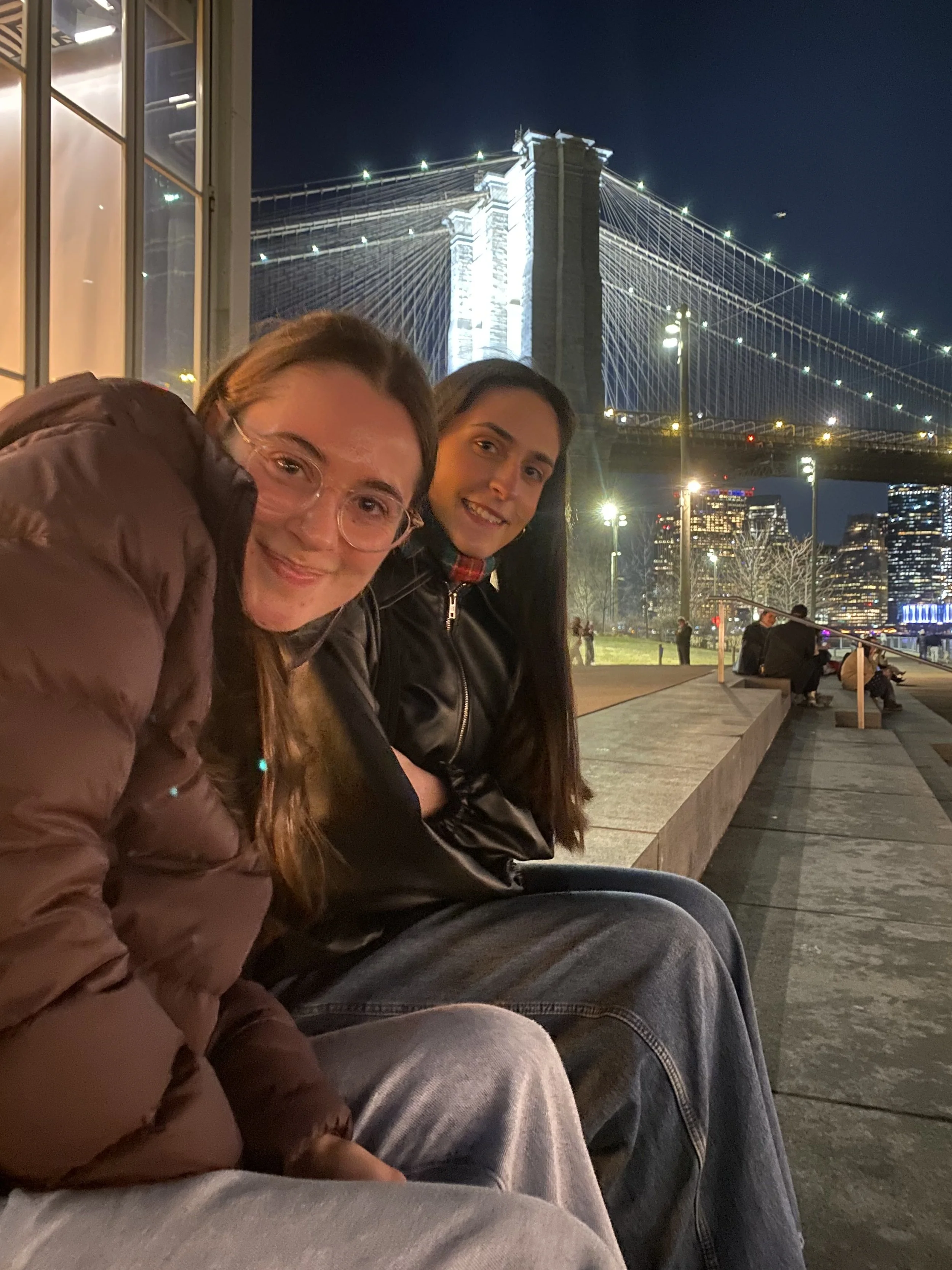 Two women sitting on a bench at night in front of the Brooklyn Bridge with illuminated towers and city buildings in the background.