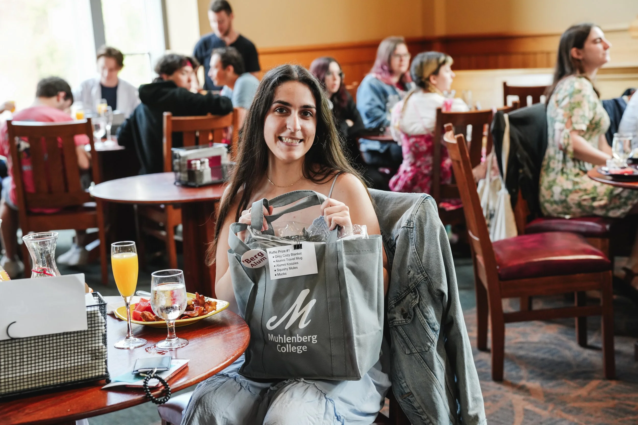 A young woman with long dark hair smiling and holding a grey college bag, sitting at a dining table during a college event, with breakfast food, drinks, and raffle prizes in front of her. Other people are seated and chatting in the background inside 