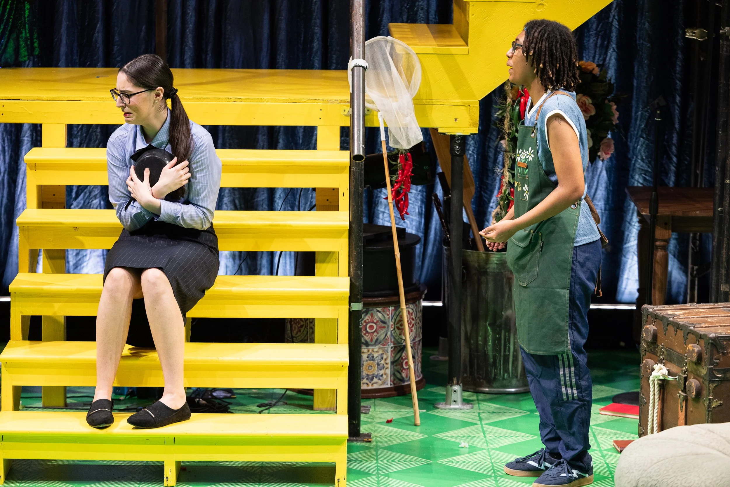 A woman with glasses, brown hair, and a blue shirt sits on yellow steps holding her chest and looking distressed, while a woman with glasses, curly hair, and a green apron stands nearby watching her.