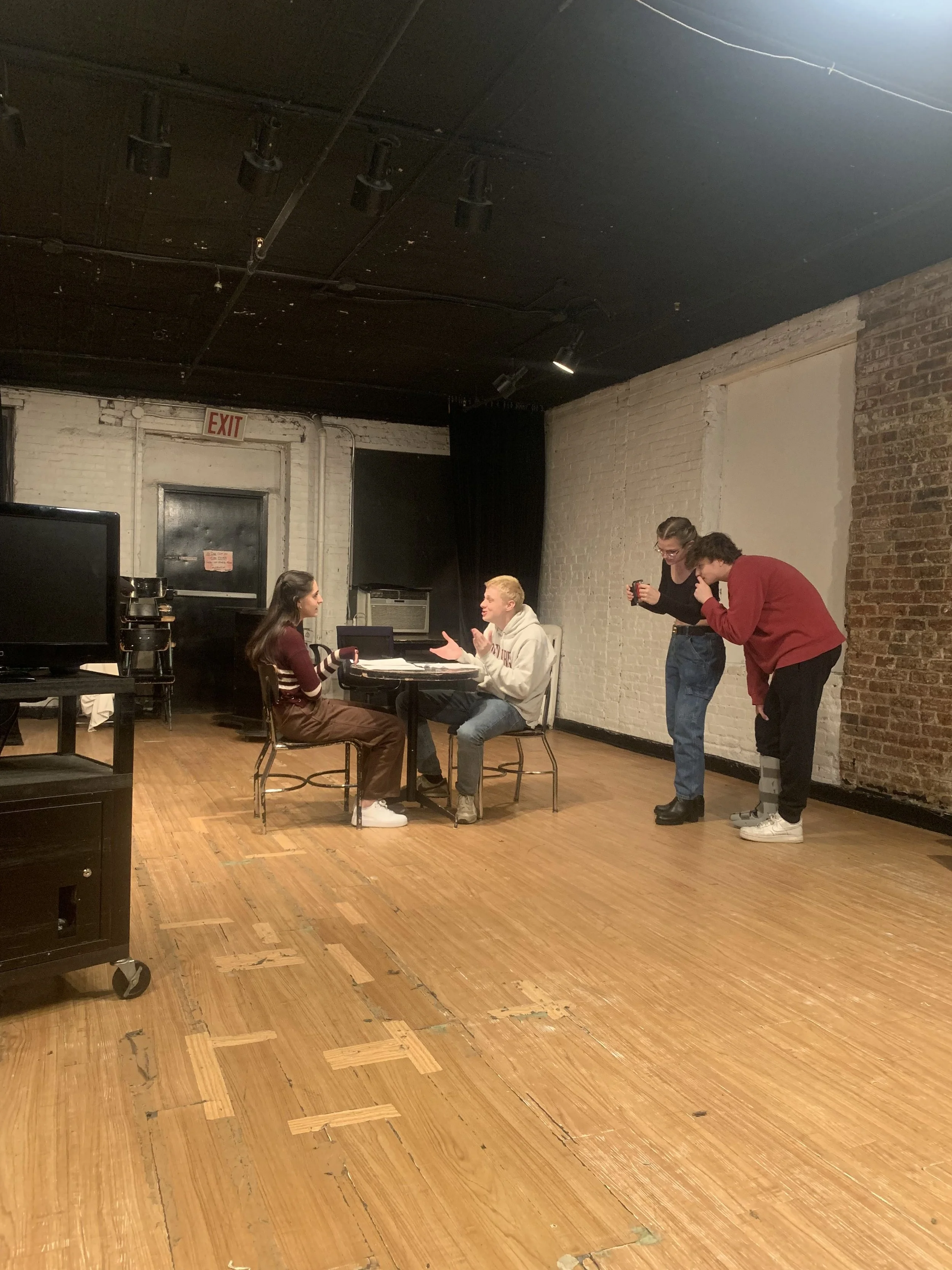 Four young people in an office or rehearsal space with exposed brick walls, engaged in conversation and humorous interaction. One person is sitting at a table with a notebook, while two others are standing and leaning towards him, smiling and sharing