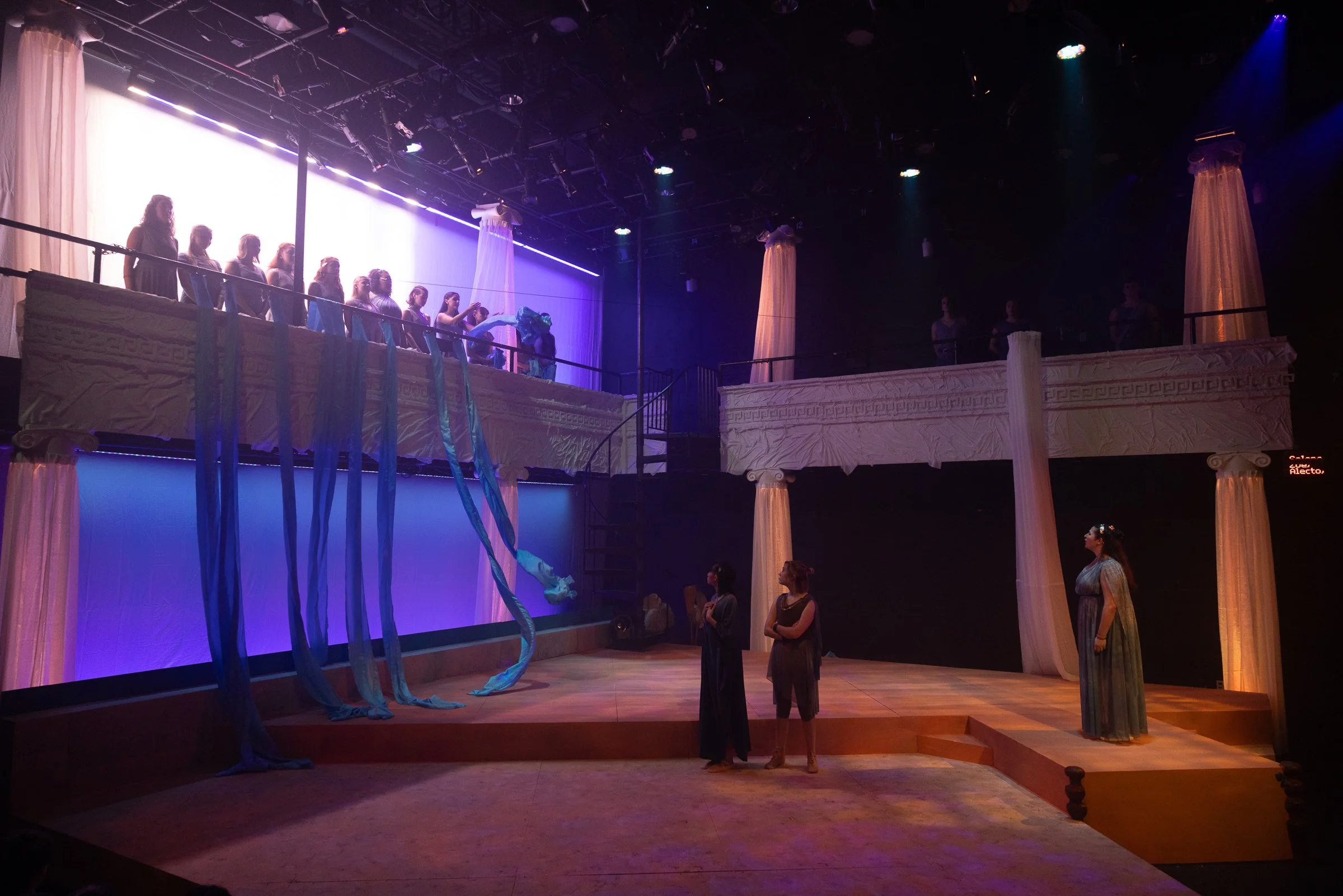 The image shows a stage set resembling ancient Greek architecture with columns and decorative elements. There is a large elevated platform with a group of people, mostly women, standing along the edge, looking towards the center. Draped fabrics and s
