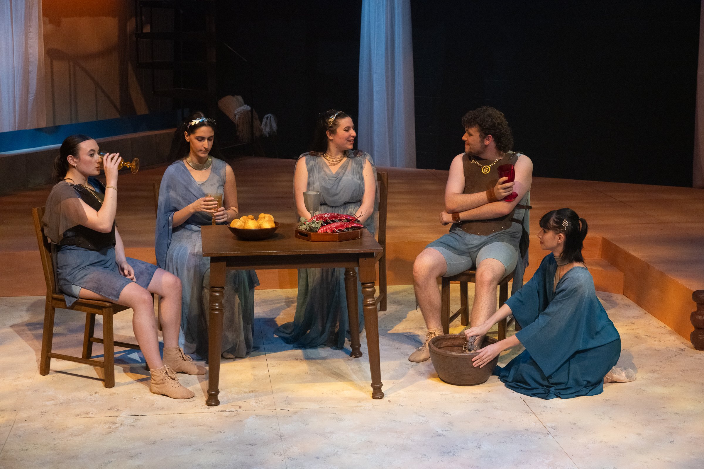 Actors in costume performing a scene on stage, with a table holding a bowl of oranges and a tray of lobster, and one actor pouring liquid from a goblet, while another actor kneels on the floor handling a basket.