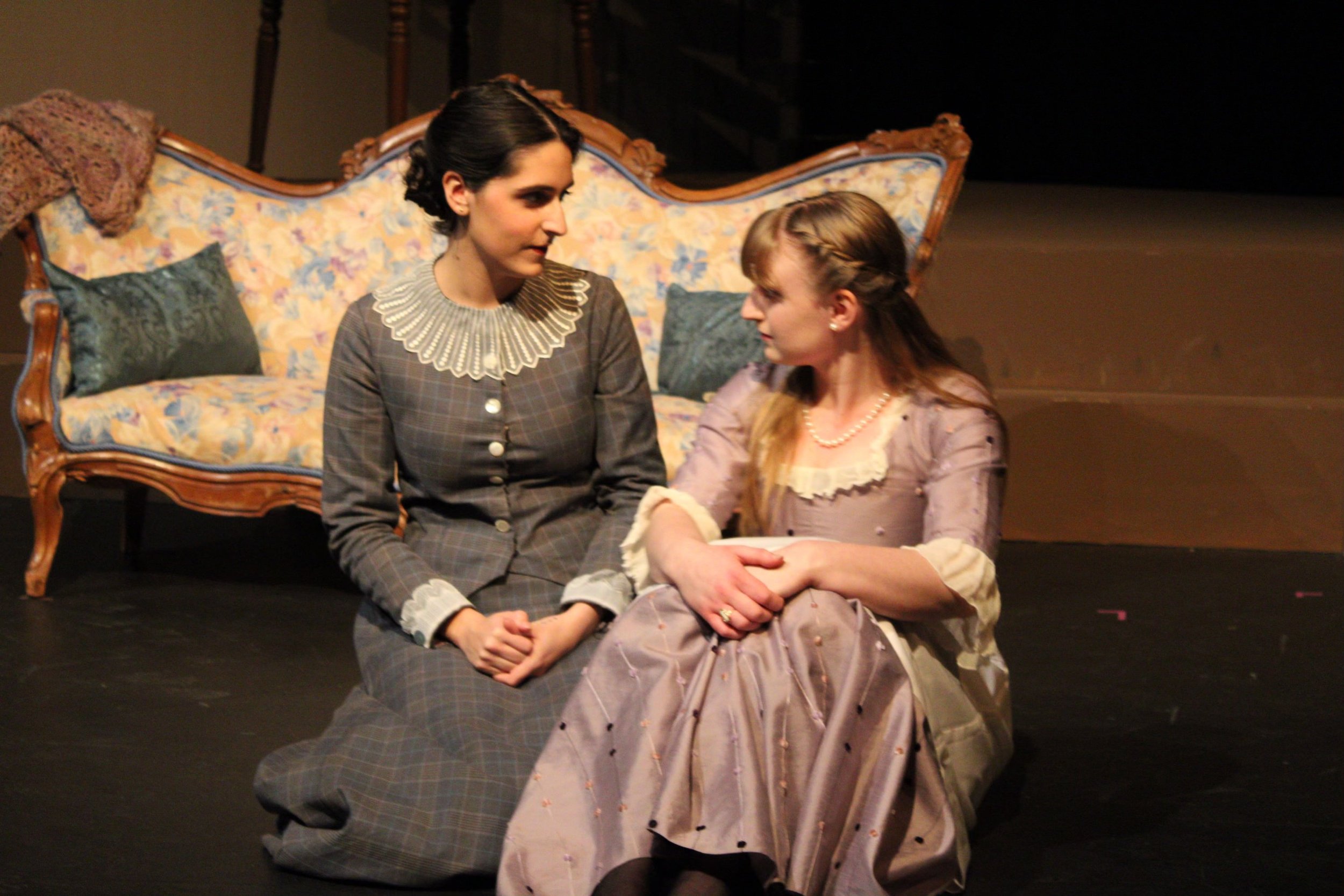 Two women sitting on the floor in front of an ornate vintage sofa, holding hands and gazing at each other. The woman on the left has dark hair and is wearing a checkered dress with a ruffled collar; the woman on the right has light hair styled with a