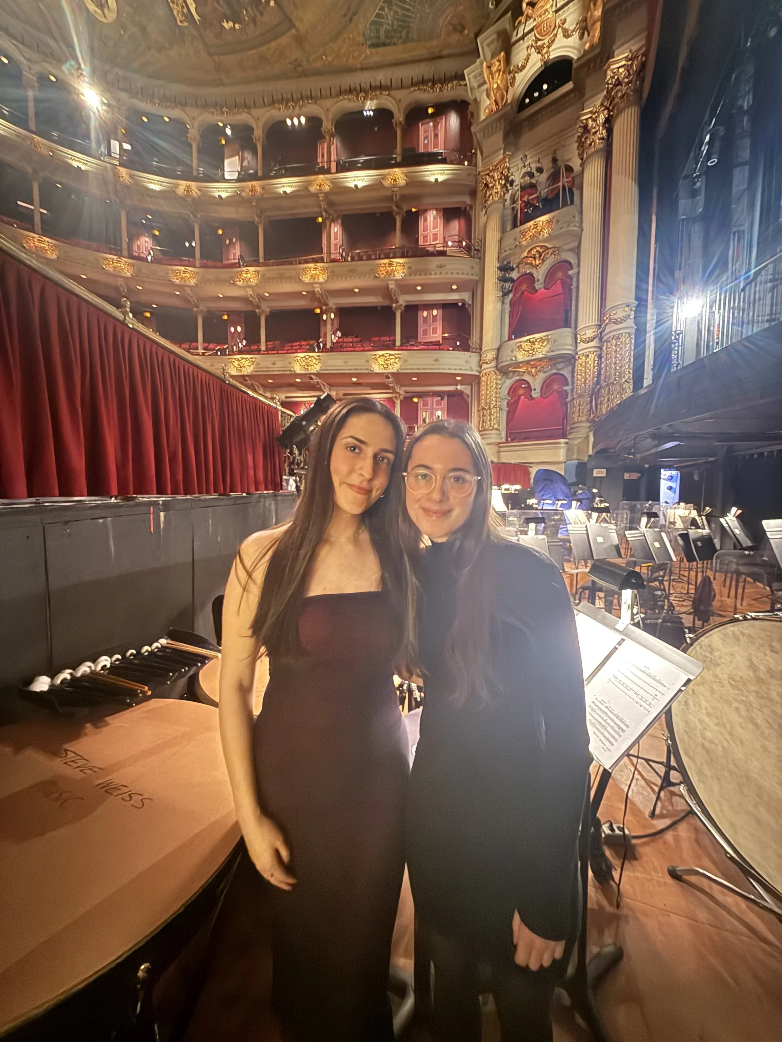 Two women standing inside a grand theater with ornate architecture, red velvet curtains, and a grand piano nearby.