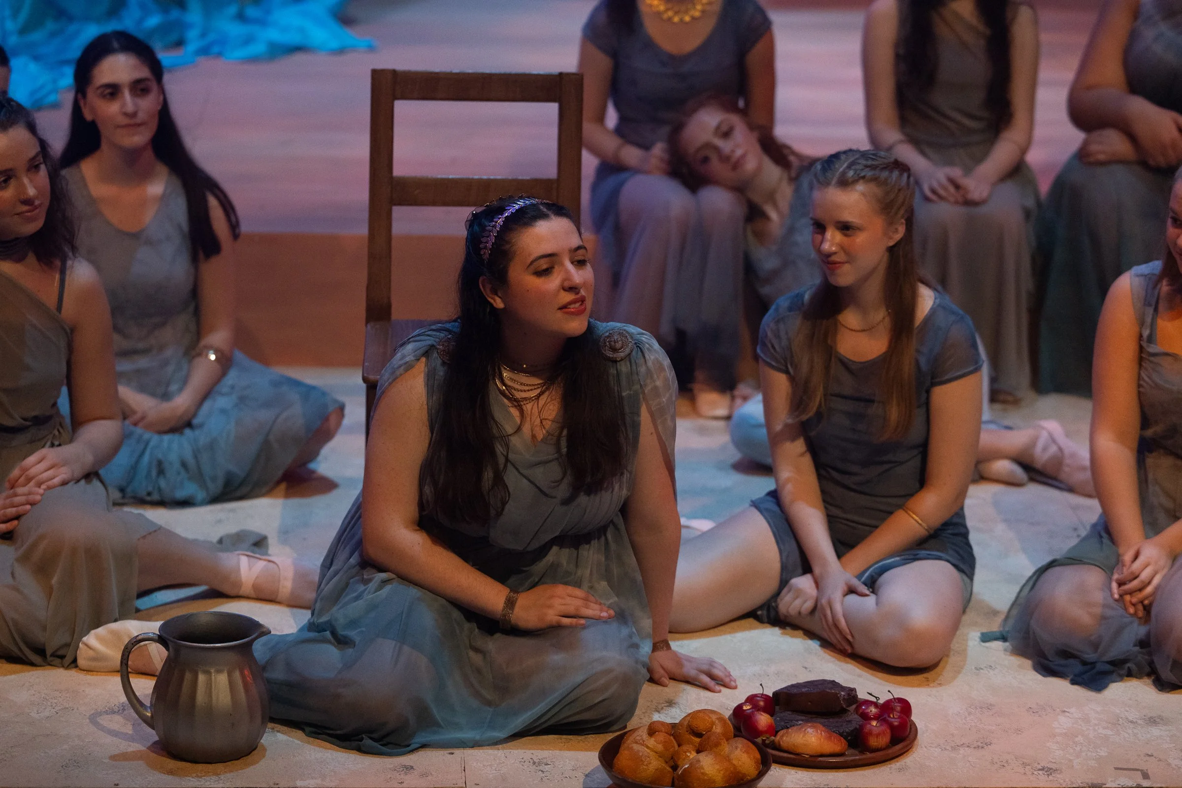 Group of women dressed in historical or traditional clothing, sitting on a stage floor with various food items including bread, nuts, apples, and a jug placed in front of them.
