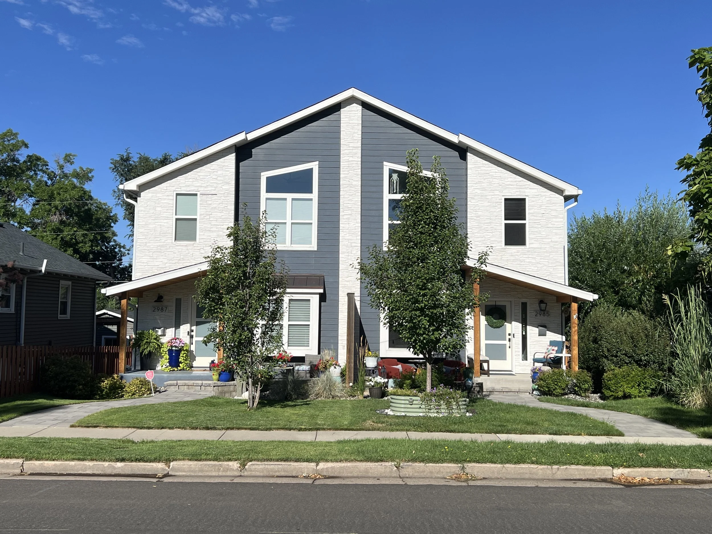 A modern two-story house with a blue and white exterior, surrounded by a well-maintained garden with trees, flowers, and a small backyard patio area. The house has several windows and a sloped roof under a bright blue sky.
