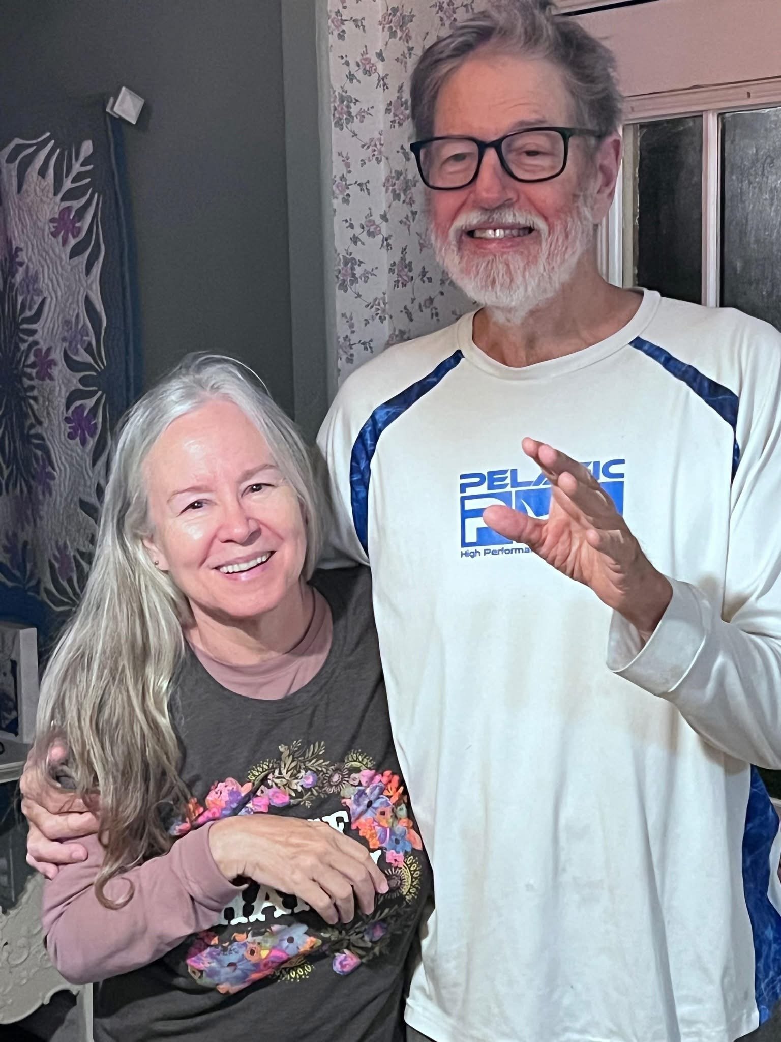 An elderly woman with long white hair, wearing a floral-patterned top and long-sleeve shirt, smiling and hugging a man dressed in a white long-sleeve shirt with a graphic logo, glasses, and gray hair, inside a room with floral wallpaper and a window.