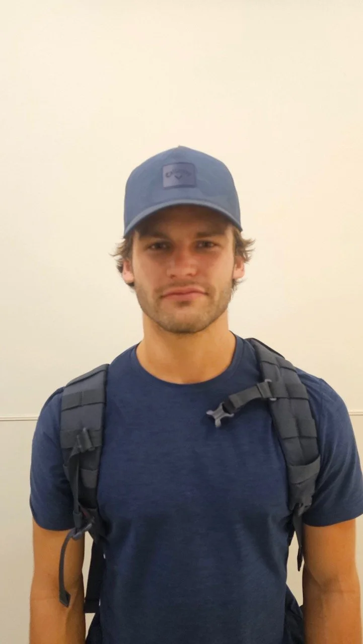Young man wearing a blue baseball cap and a navy blue t-shirt, carrying a backpack, standing against a plain beige wall.