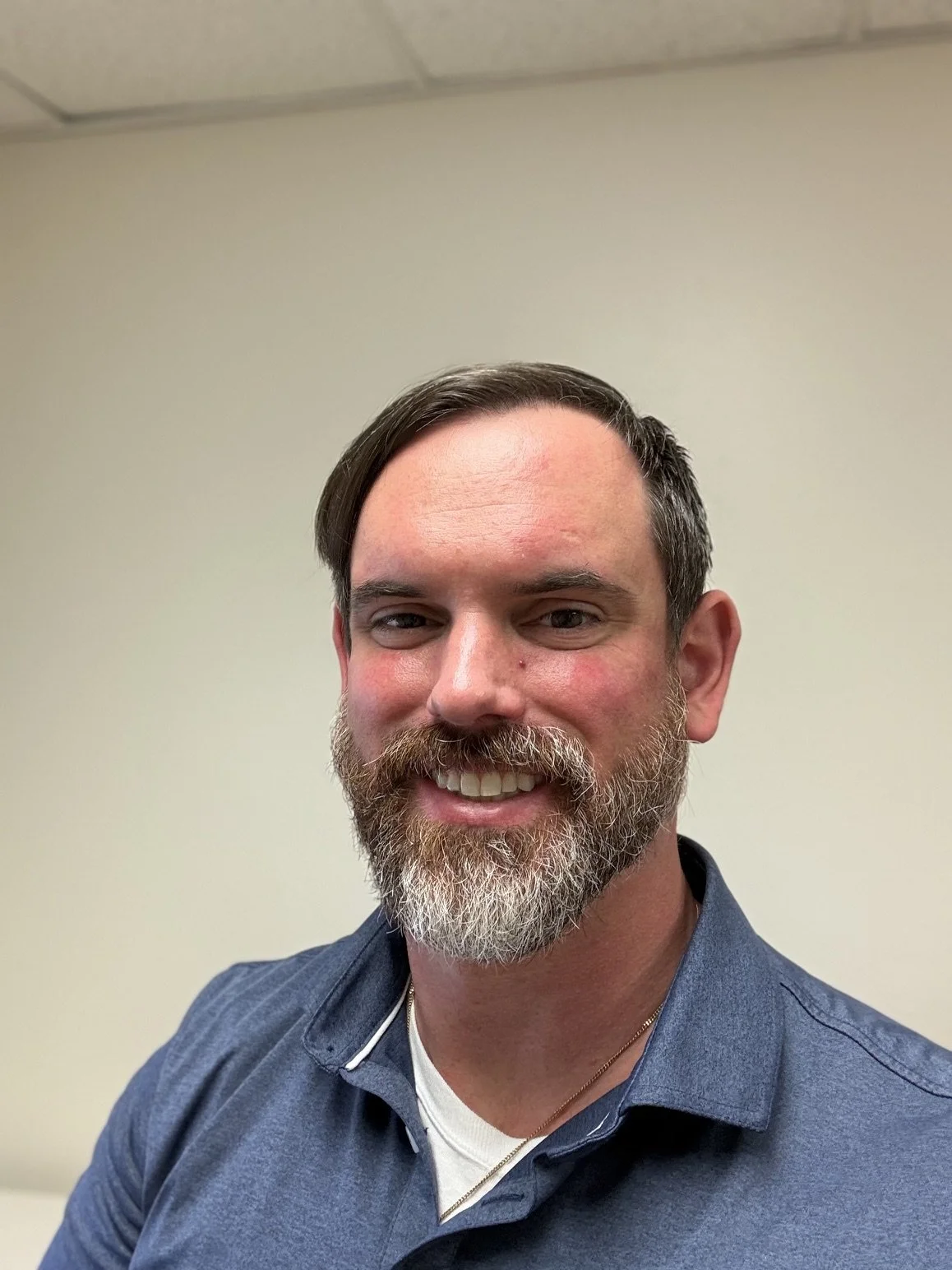 A smiling man with a beard and mustache, wearing a collared shirt, posing indoors against a plain wall.