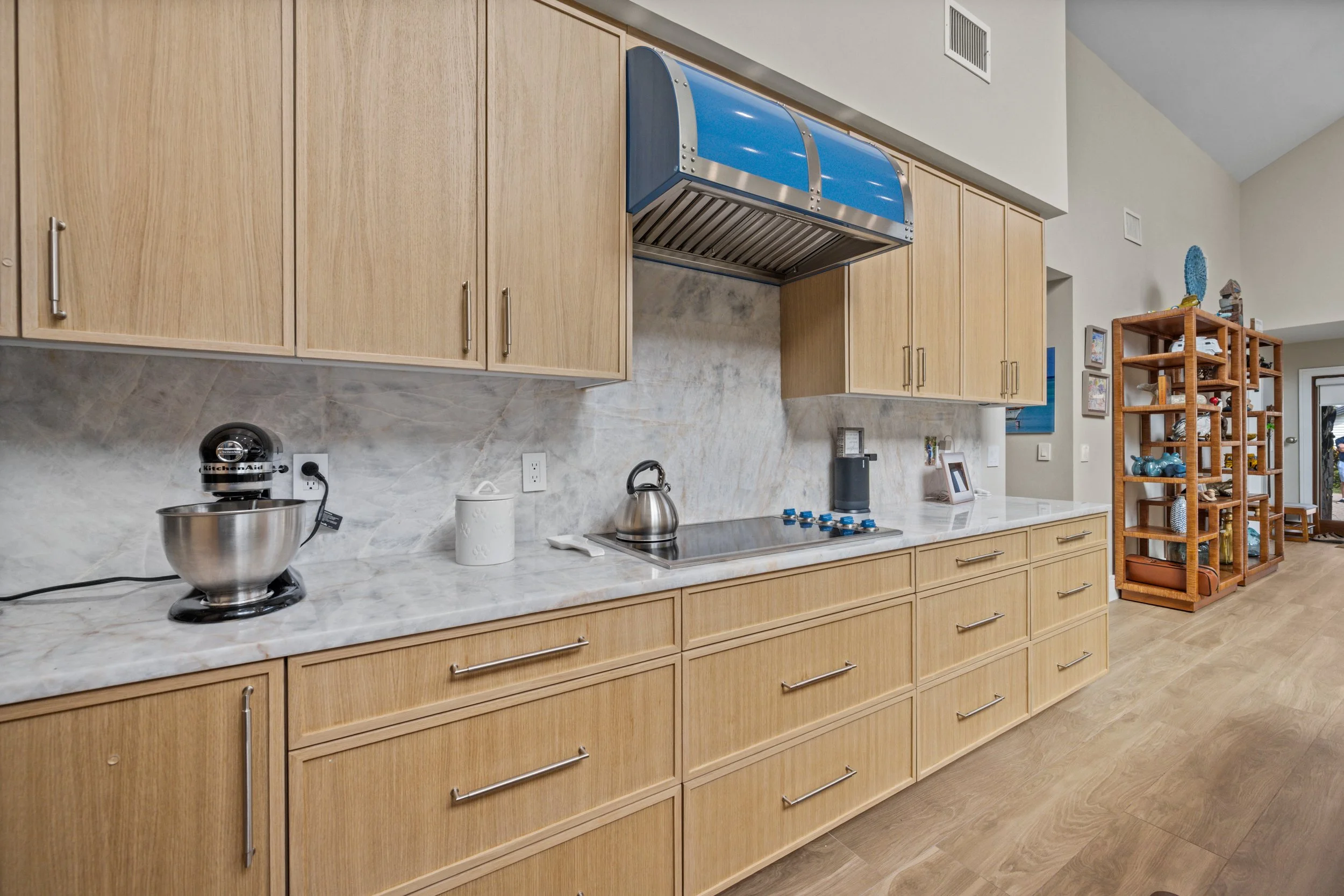 A kitchen with wooden cabinets, marble countertops, a stainless steel kettle, a KitchenAid mixer, and a wooden shelving unit with various decor and kitchen items.