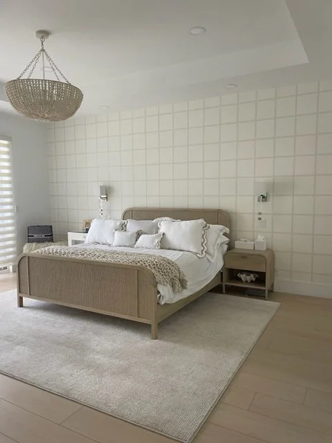 Minimalist bedroom with a beige bed, white bedding, and pillows, two white nightstands with lamps, a chandelier, and a light-colored rug on wooden floor.