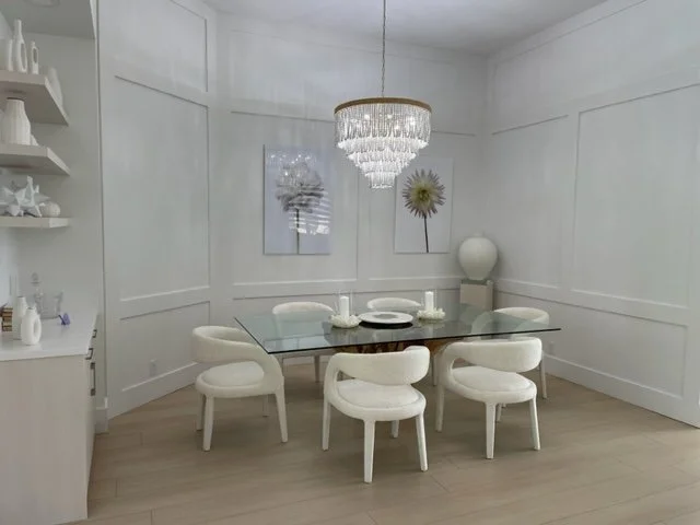 Modern dining room with white walls, a glass table, six white upholstered chairs, a crystal chandelier, and decorative art on the wall.