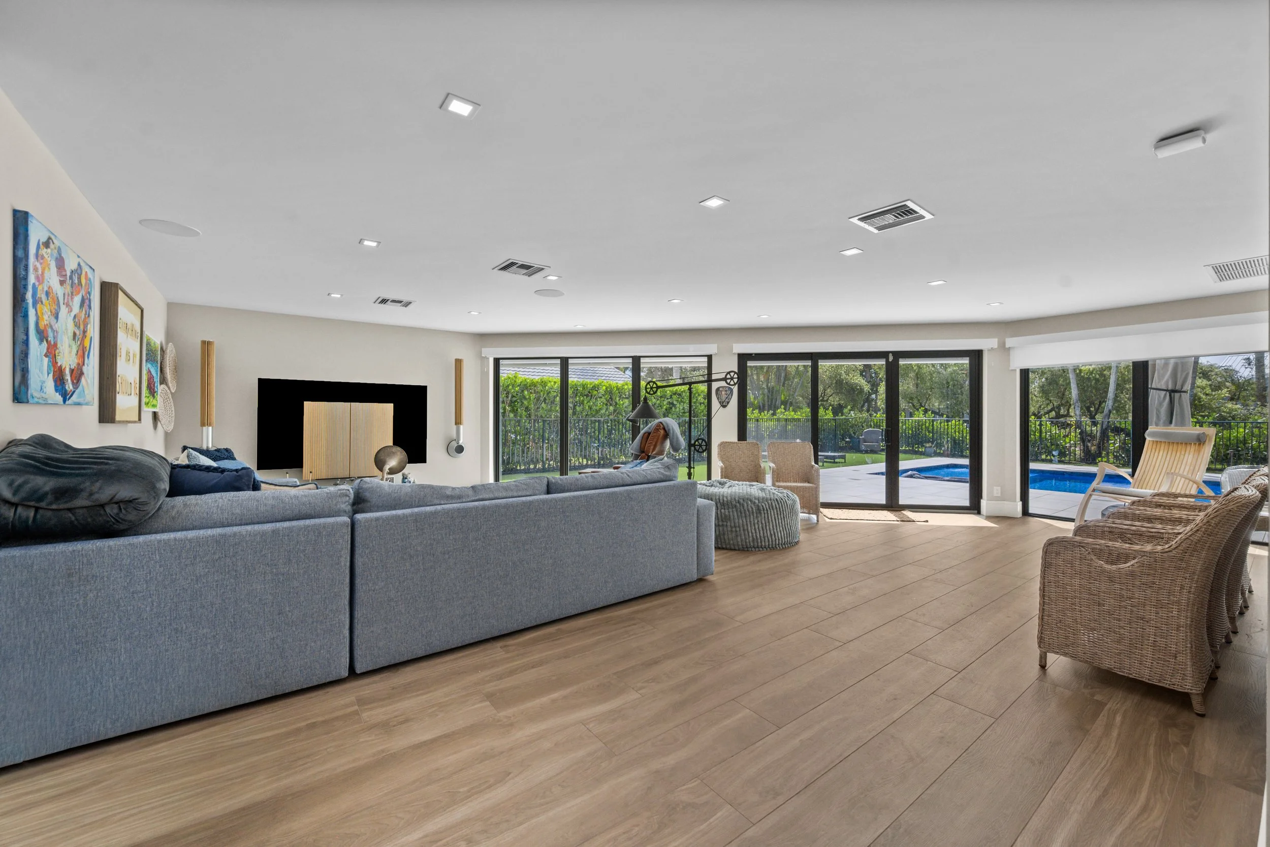 Spacious living room with large sliding glass doors leading to a backyard with pool, featuring a gray sectional sofa, wicker chairs, colorful artwork on the wall, and wood-like flooring.