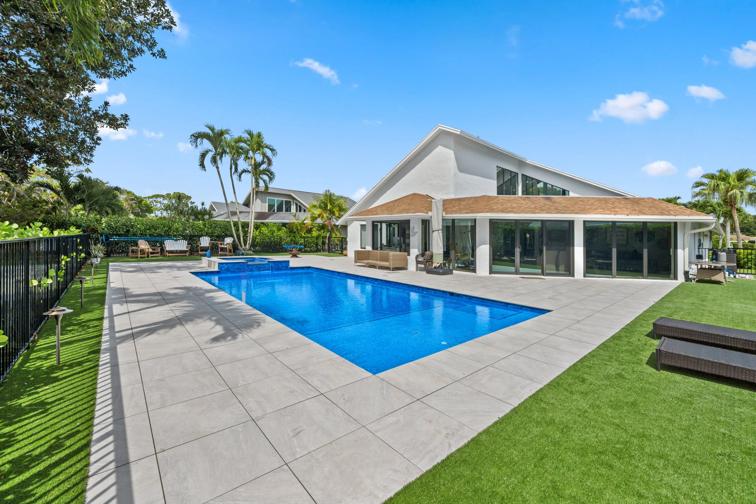 A backyard with a swimming pool, surrounded by tan tiles, with outdoor furniture and lush green trees and grass, and a modern white house with large windows under a blue sky with some clouds.