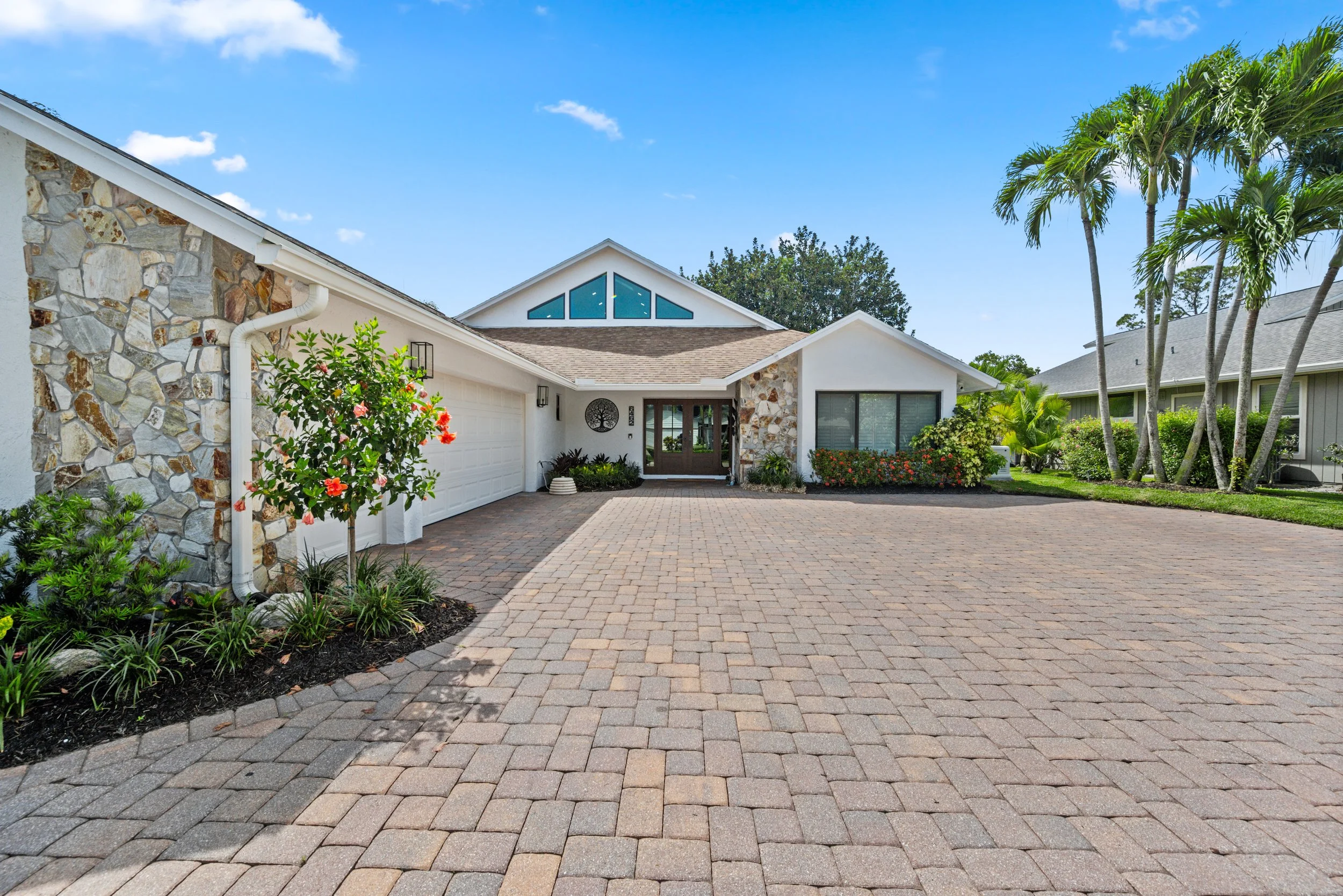Front view of a modern house with a stone and white exterior, a driveway paved with brick, surrounded by lush greenery and tall palm trees, under a blue sky with scattered clouds.